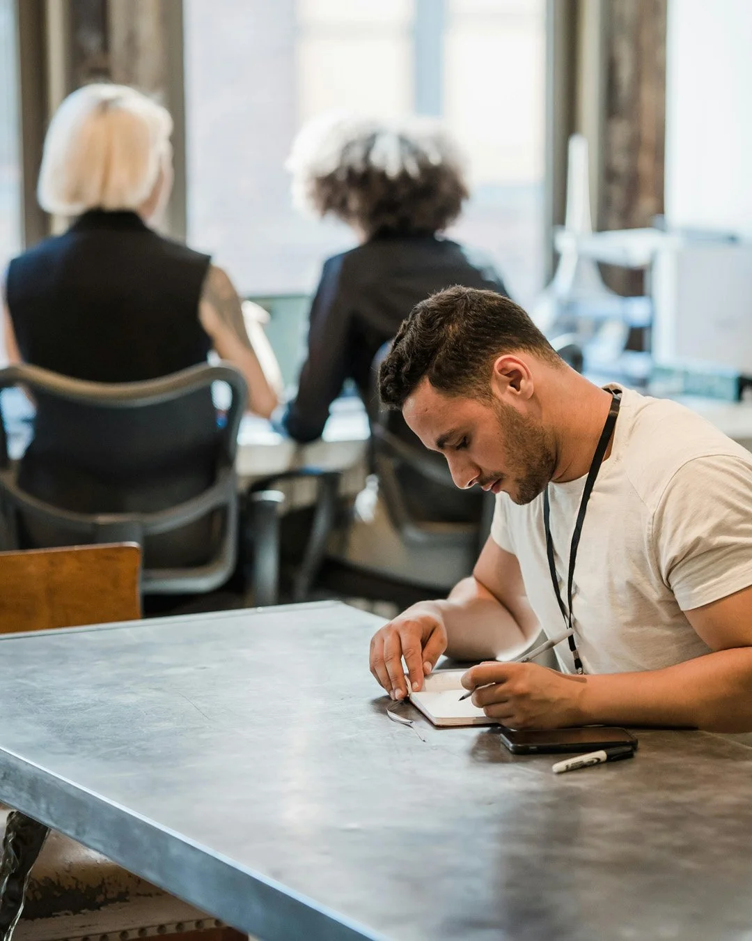 Young man taking notes at a desk in a busy office with three women working in the background.