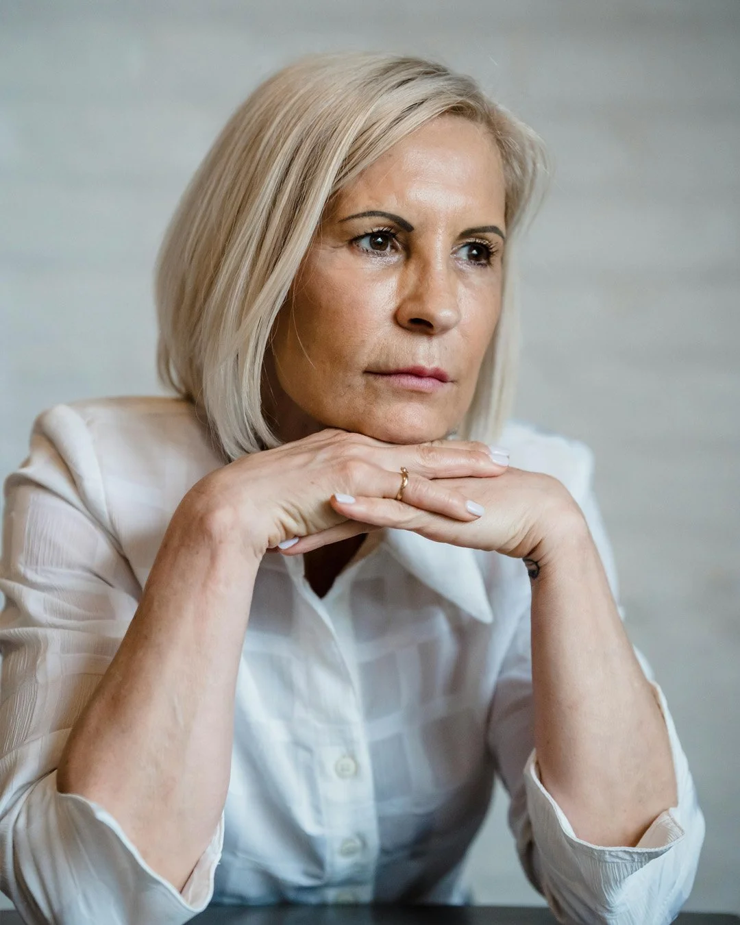 A middle-aged woman with blonde hair sitting with her chin resting on her hands, looking thoughtful.