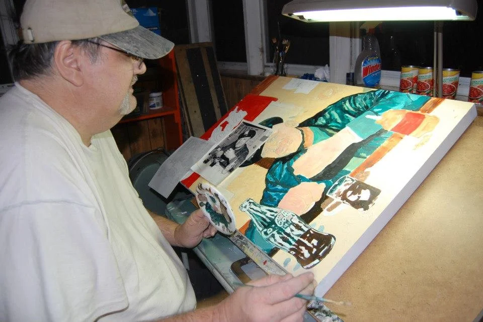 An artist in a beige cap and glasses painting a portrait on a canvas, with a palette in hand, in a well-lit room with art supplies and reference photos on the table.