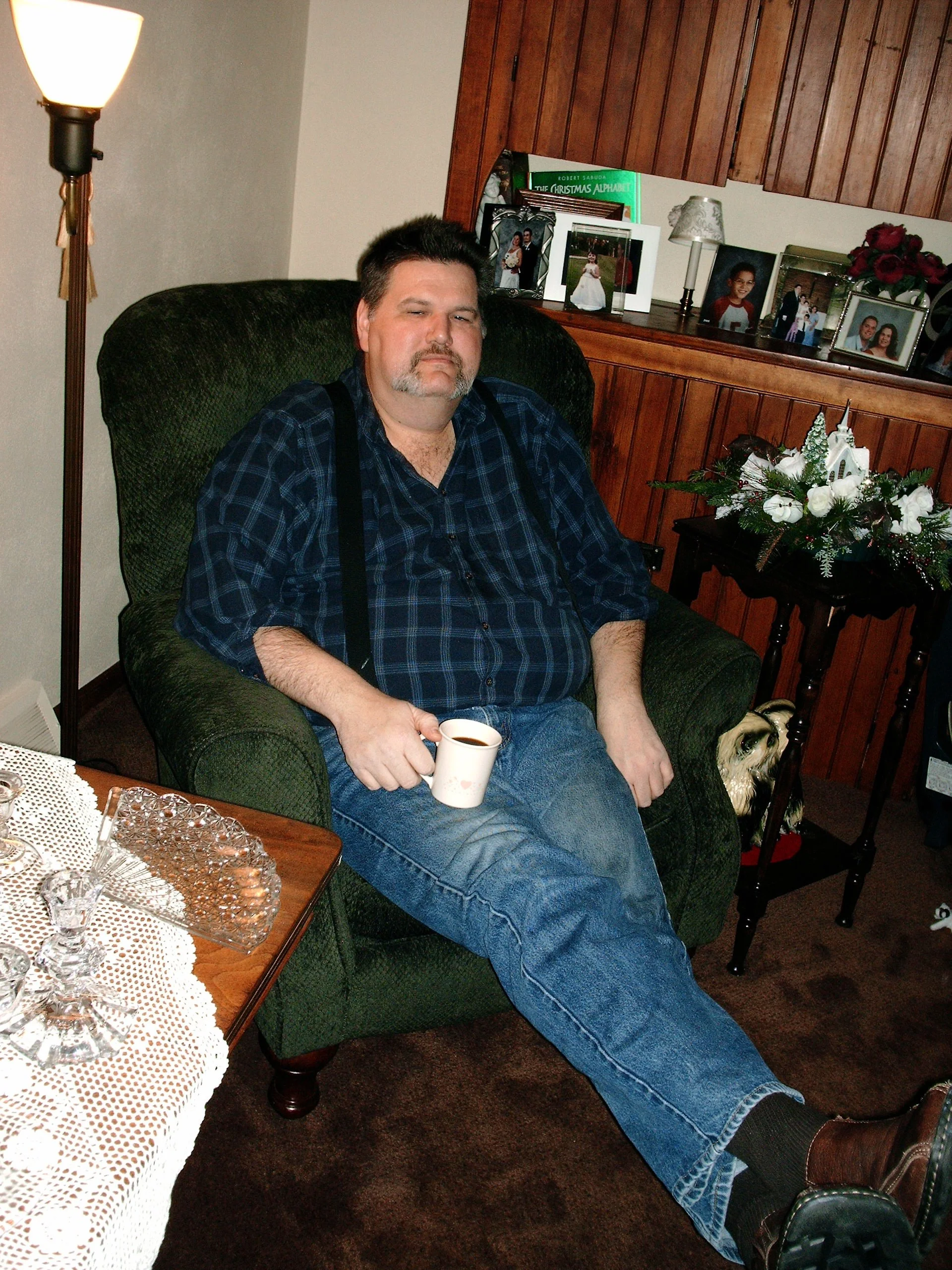 A man sitting on a green armchair in a room decorated with family photographs, a bouquet of flowers, and Christmas decor, holding a coffee cup.