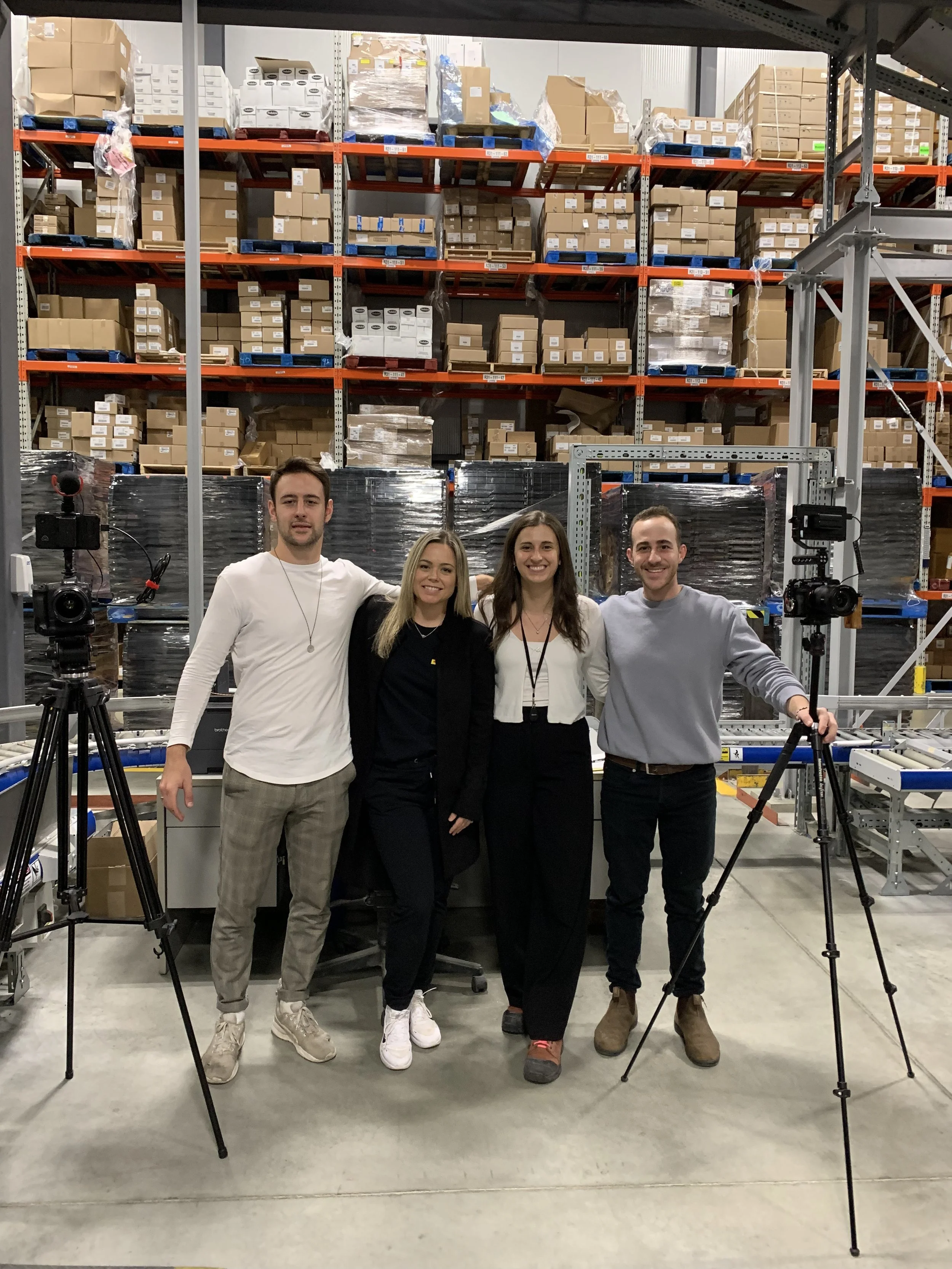 Group of four people standing in a warehouse, with shelves filled with boxes behind them, and camera equipment set up to take a group photo.