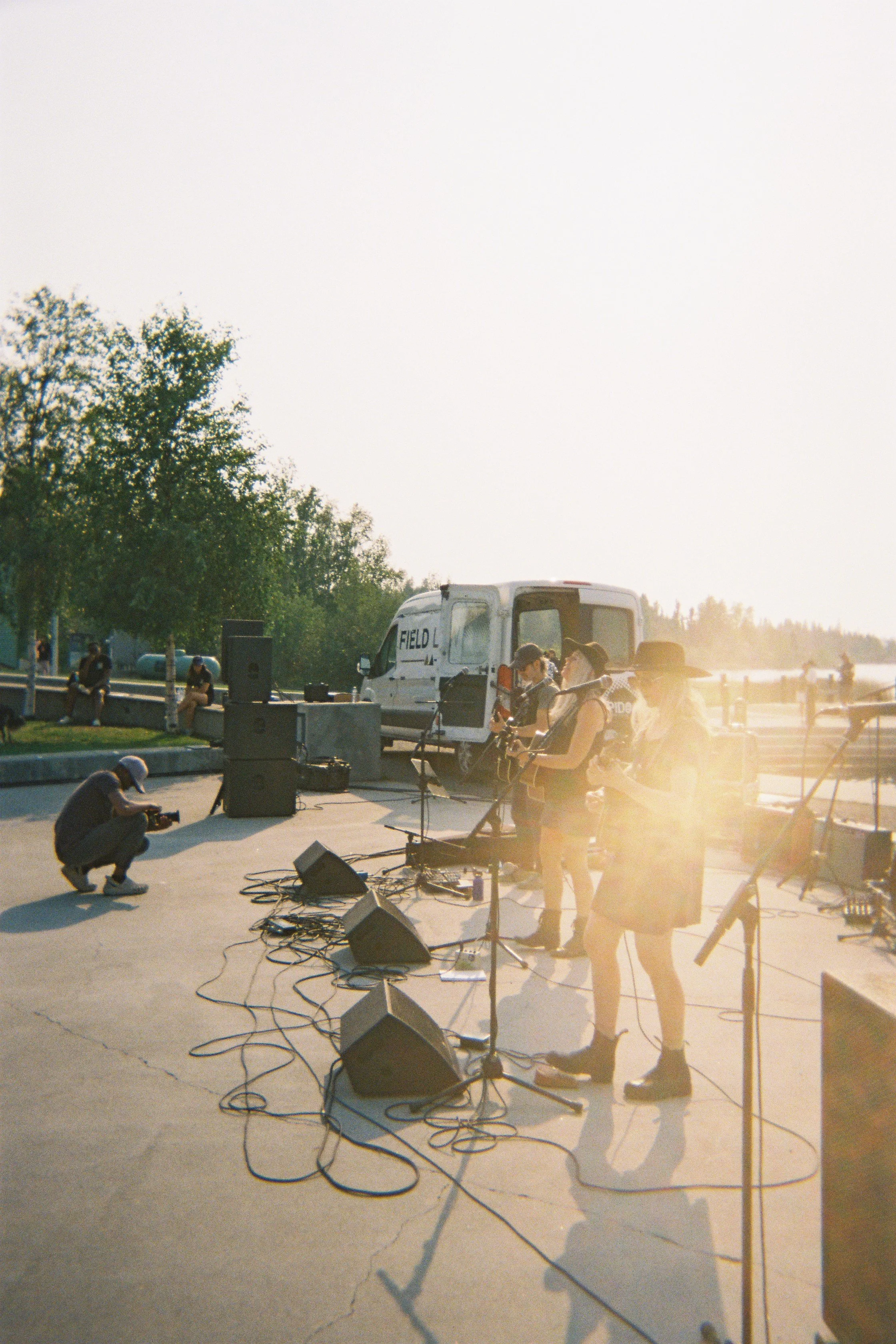 People setting up musical equipment outdoors during sunset, with a van, trees, and a few onlookers nearby.