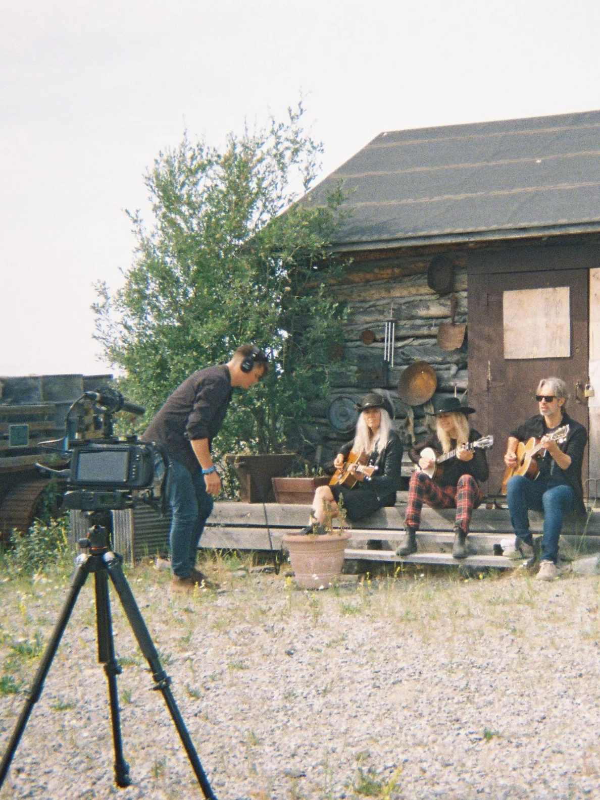 Three musicians playing guitars outdoors on a porch as a person adjusts camera equipment nearby during an outdoor filming session.