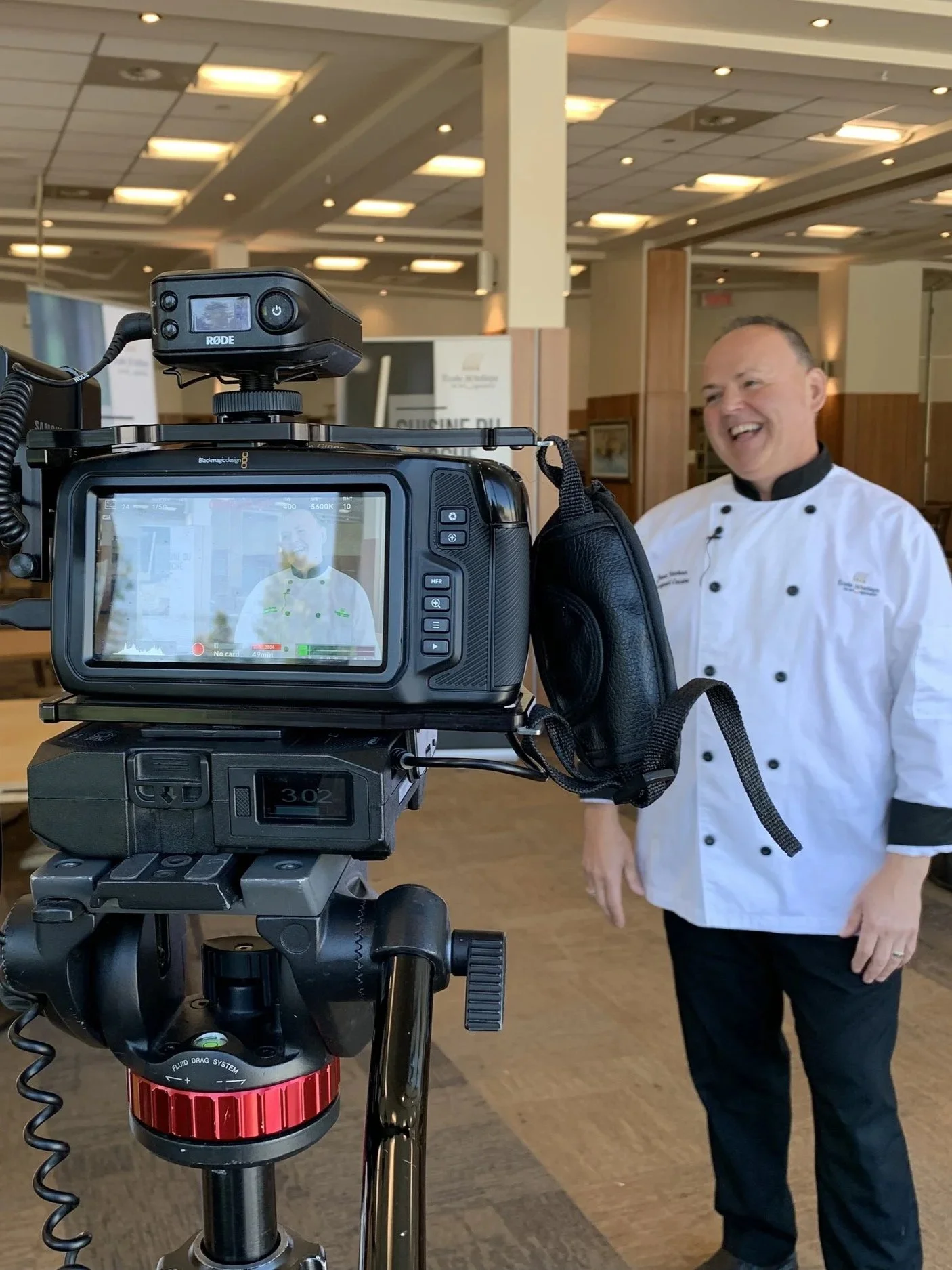 A chef in a white uniform with black buttons is smiling and standing behind a professional video camera that is recording him. The scene is inside a well-lit, modern room with wooden accents.