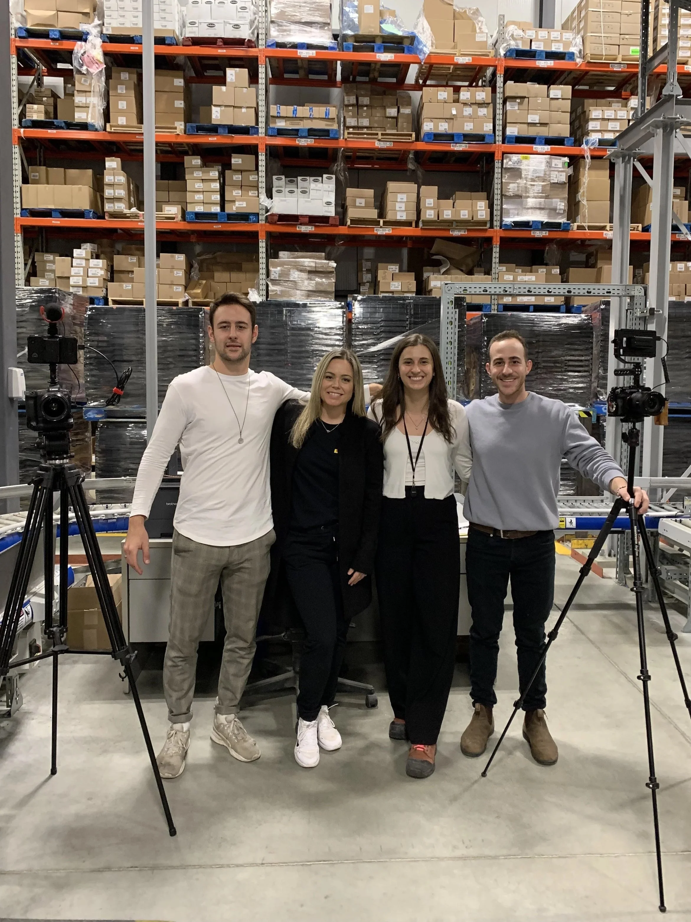 Four young adults standing together in a warehouse, smiling at the camera. They are flanked by tripods with cameras, and there are shelves filled with cardboard boxes and pallets of supplies in the background.