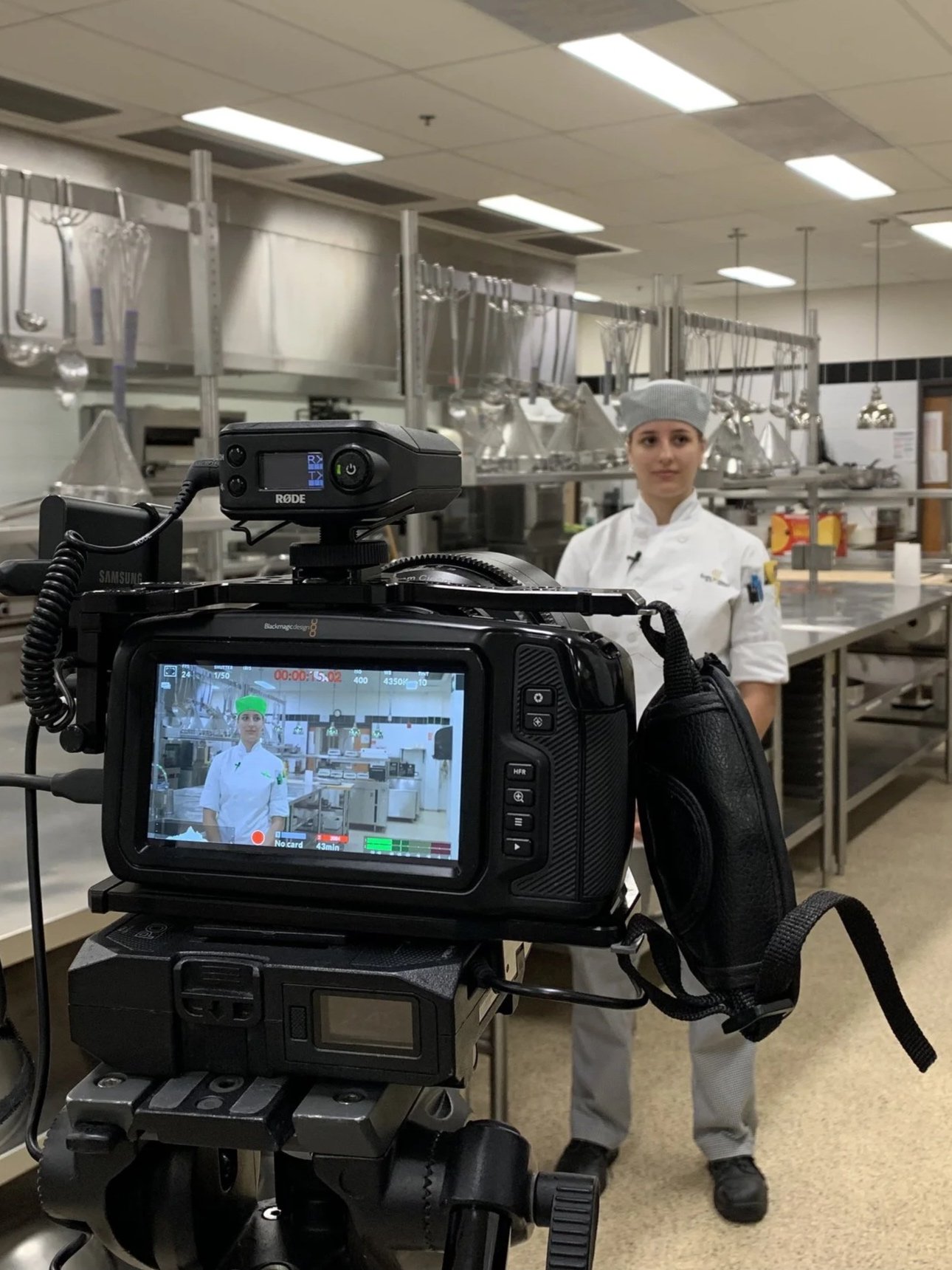 A professional video camera recording a female chef in a commercial kitchen. The chef is wearing a gray hairnet, a white chef's coat, and dark footwear. The kitchen background includes metallic shelves filled with kitchen tools and equipment, and the ceiling has bright fluorescent lights.