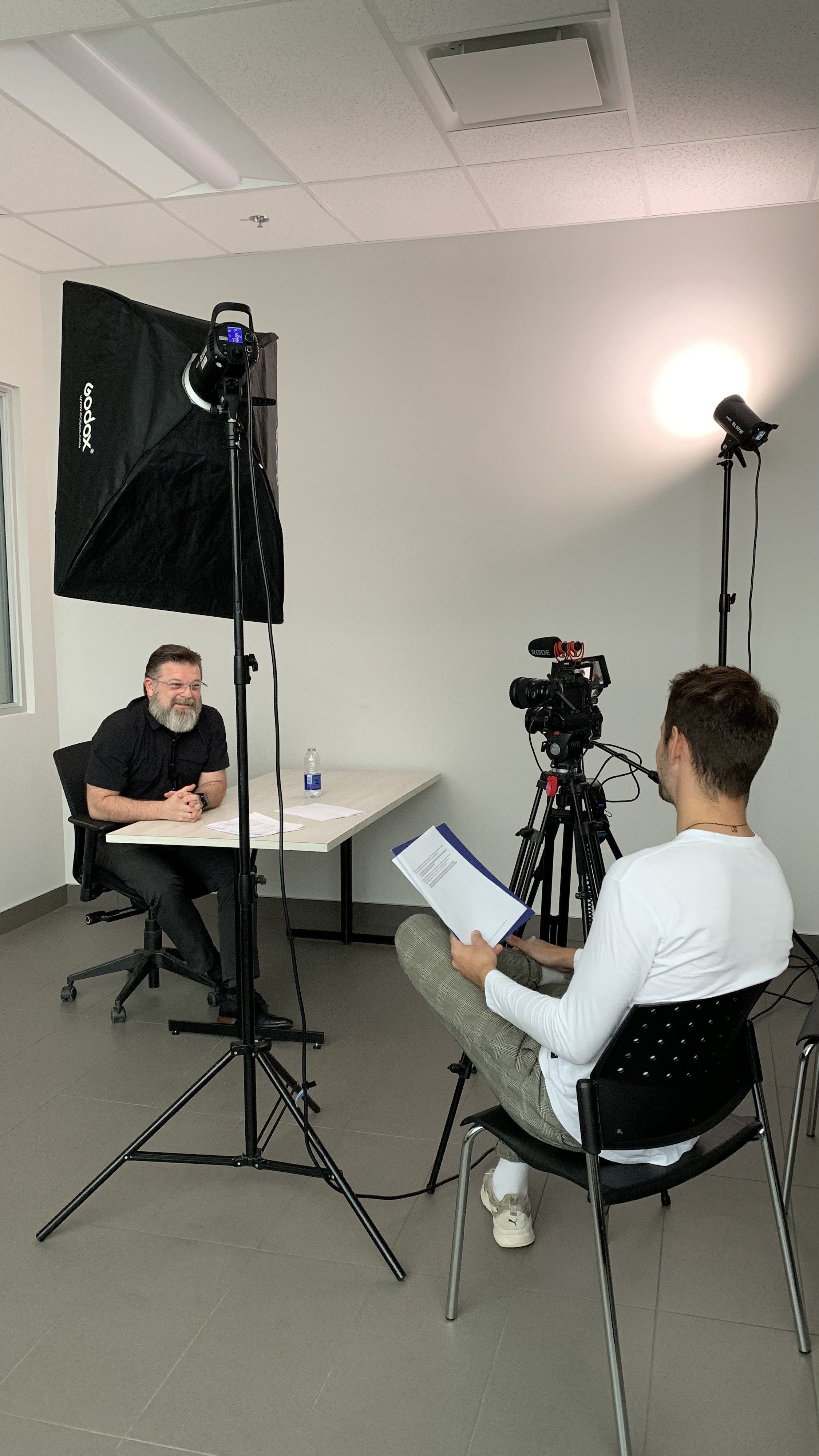 A man with a beard and glasses is sitting at a table in front of a camera during an interview or recording session, with studio lights and a camera operator taking notes.