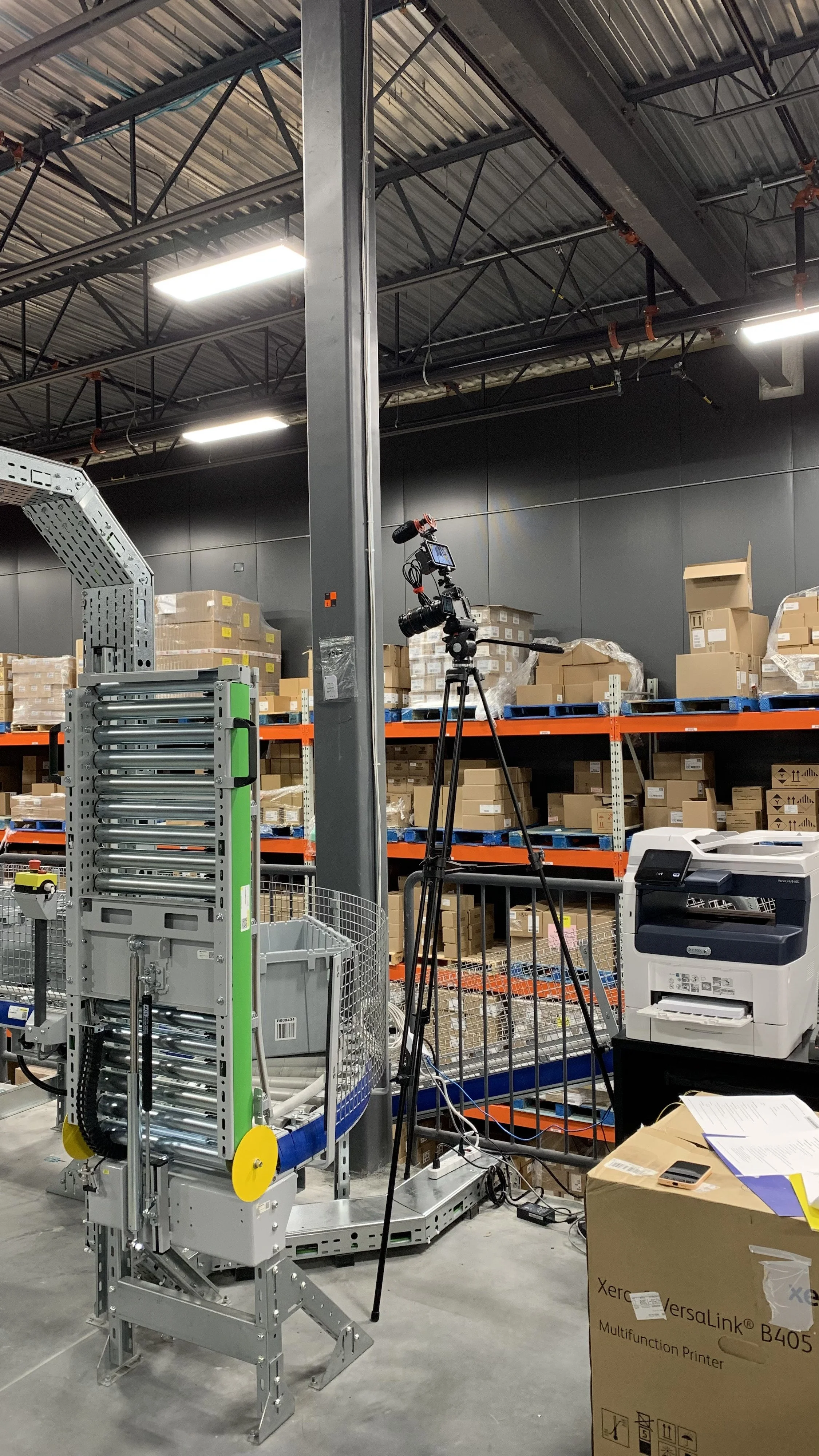 A robot with metal rollers and a yellow wheel in a warehouse with shelves filled with boxes and crates, camera on a tripod, industrial printer, and various office supplies.
