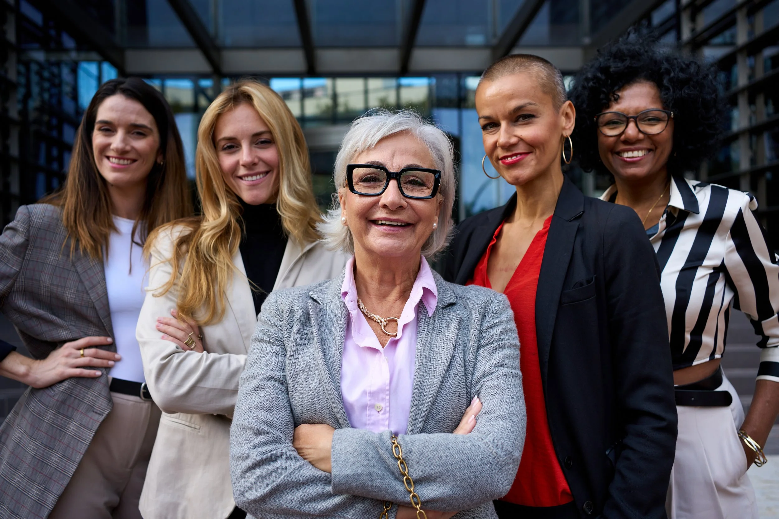 Group of five women standing outside in front of a modern glass building, smiling and posing for the photo. The woman in the center has short gray hair and glasses, wearing a gray blazer and pink shirt.