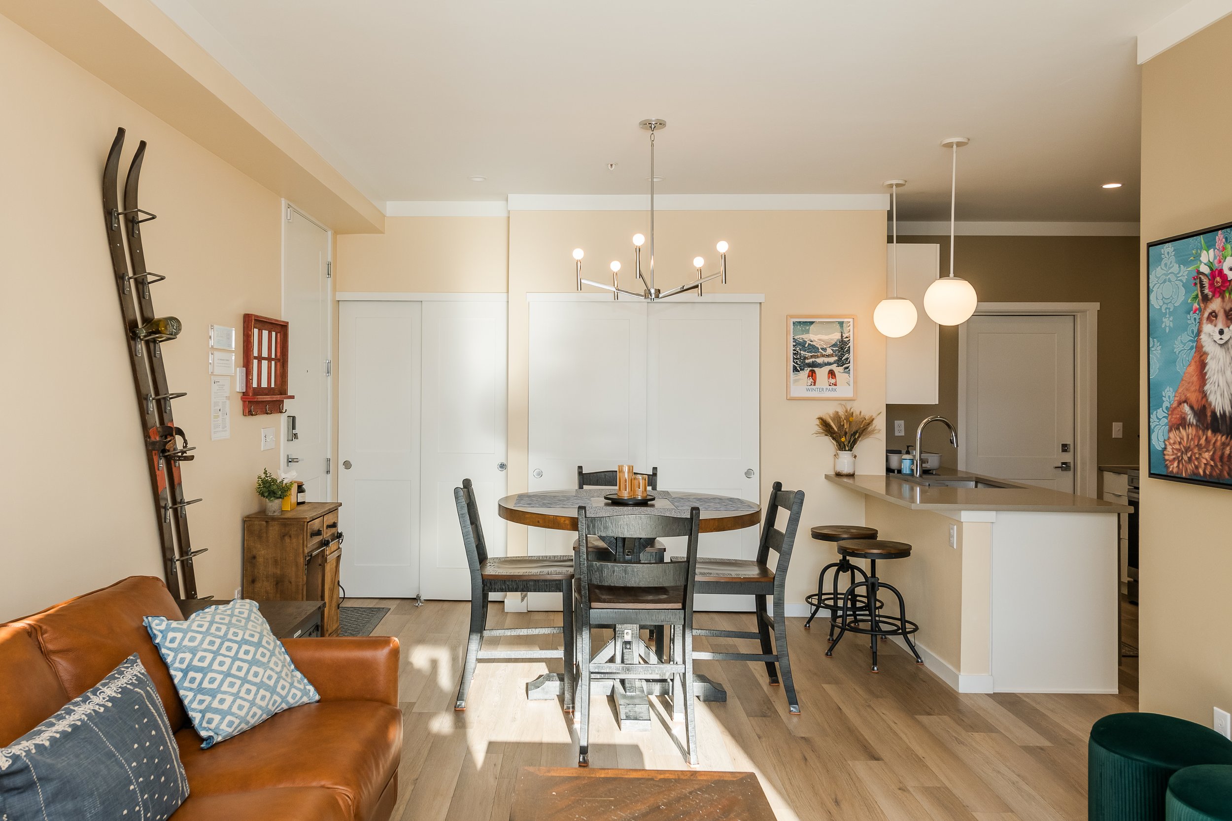A dining area and kitchen in a home with beige walls and wooden flooring. There is a round dining table with six chairs, a hanging modern chandelier, and two hanging round pendant lights. Decor includes framed artwork, a vase with dried plants, and a