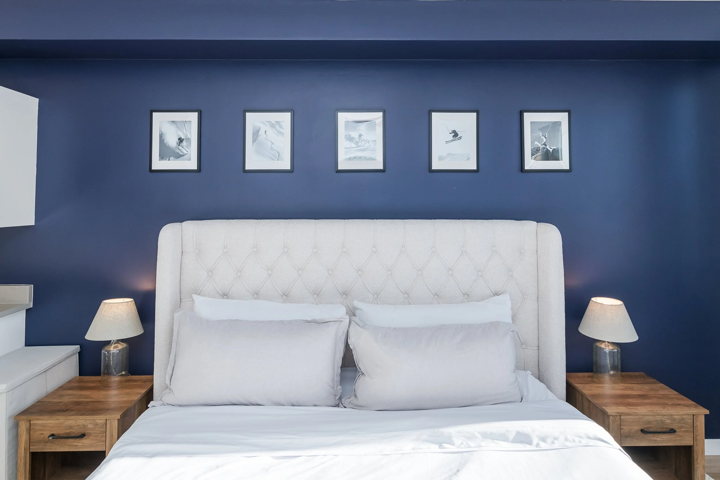 A bedroom with a white upholstered headboard, white bedding, and two matching nightstands with lamps. The wall behind the bed is painted dark blue and features five framed black-and-white photographs of winter sports.