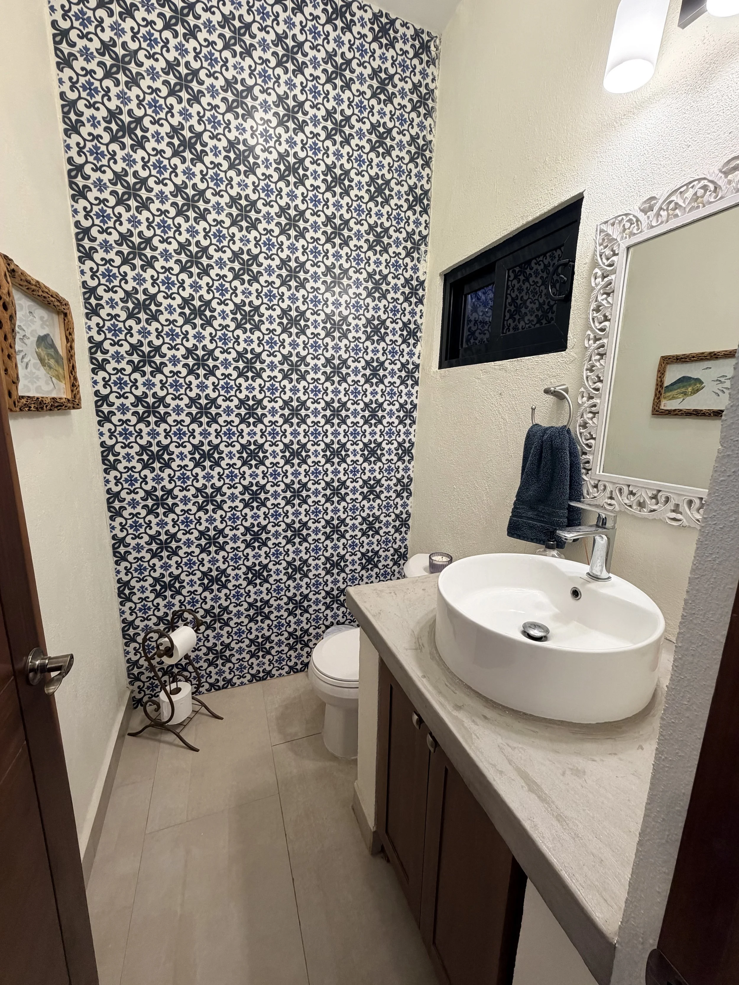 A small bathroom with a white oval sink on a beige marble countertop, a wall mirror with an ornate white frame, a dark towel hanging on a silver hook, a black window with a view of a patterned blue and white tiled wall in the shower area, and two toi