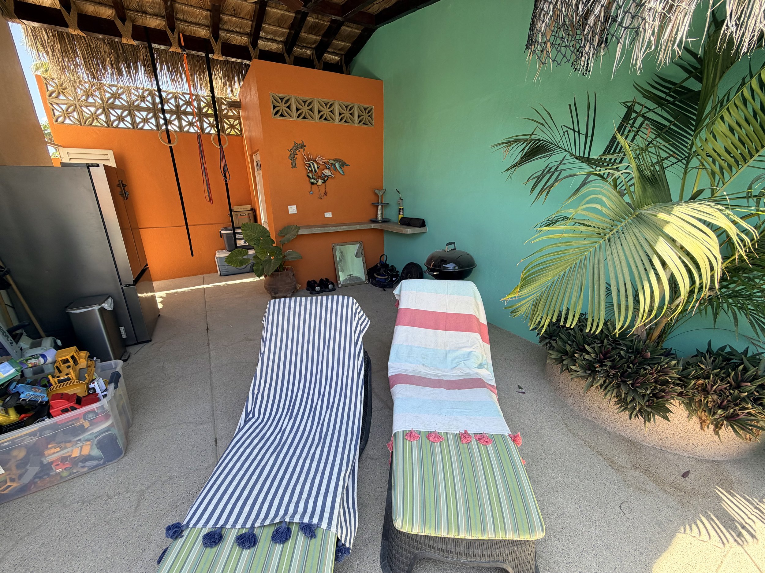 Outdoor patio with colorful walls and tropical plants, featuring striped couches, a table with a striped tablecloth, a barbecue grill, and various toys and decorations.