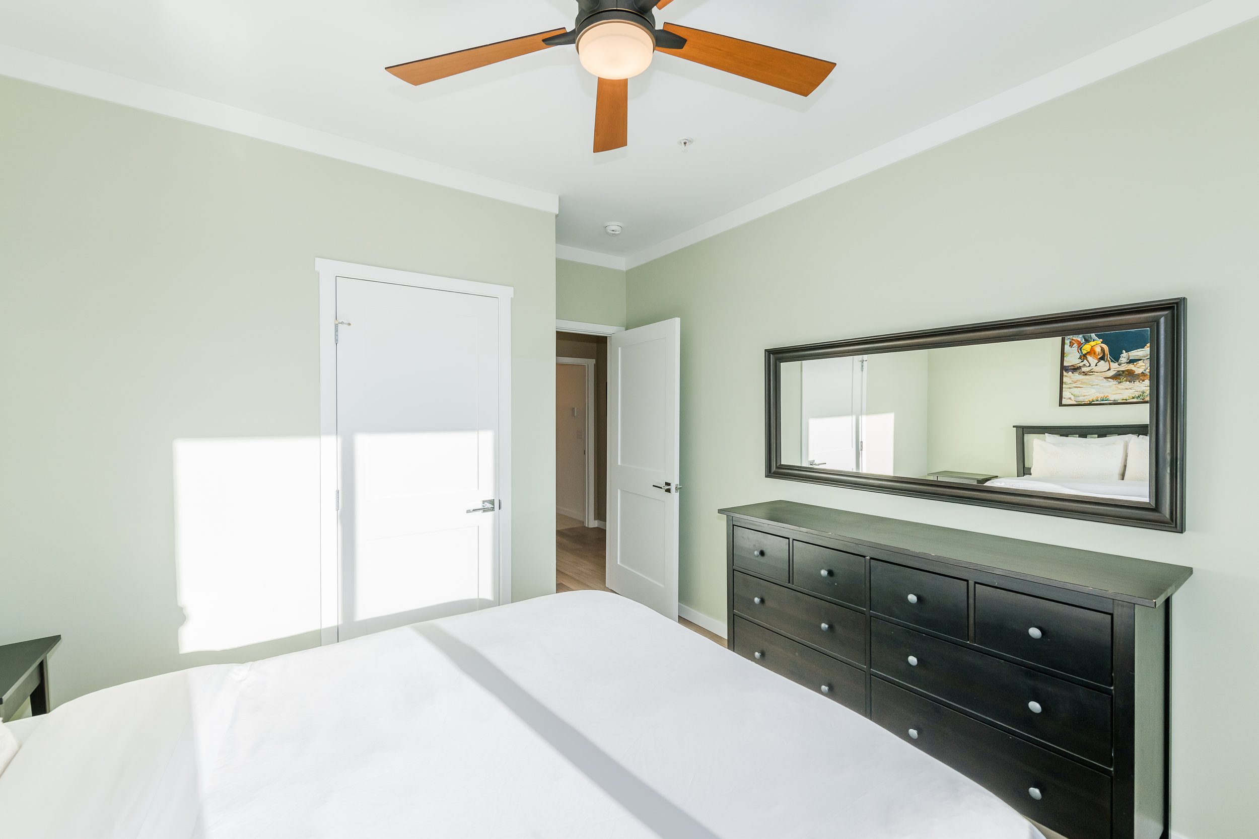 A bedroom with light green walls, a large mirror, a dark dresser, a ceiling fan with wooden blades, and framed artwork depicting horses.