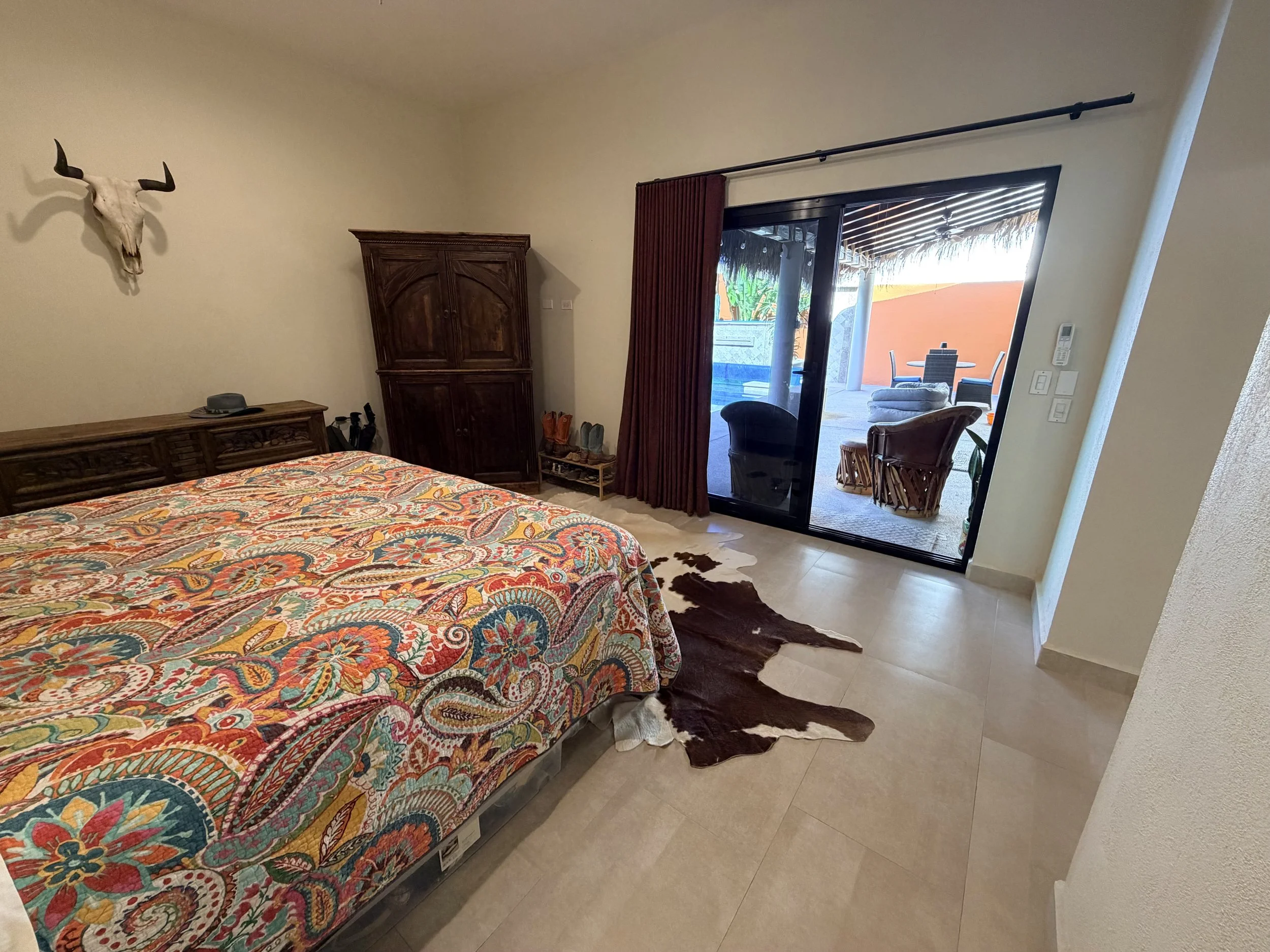 A bedroom with a bed covered in a colorful, intricate patterned bedspread, a wooden wardrobe, and a sliding glass door leading to an outdoor patio with seating and a thatched roof.