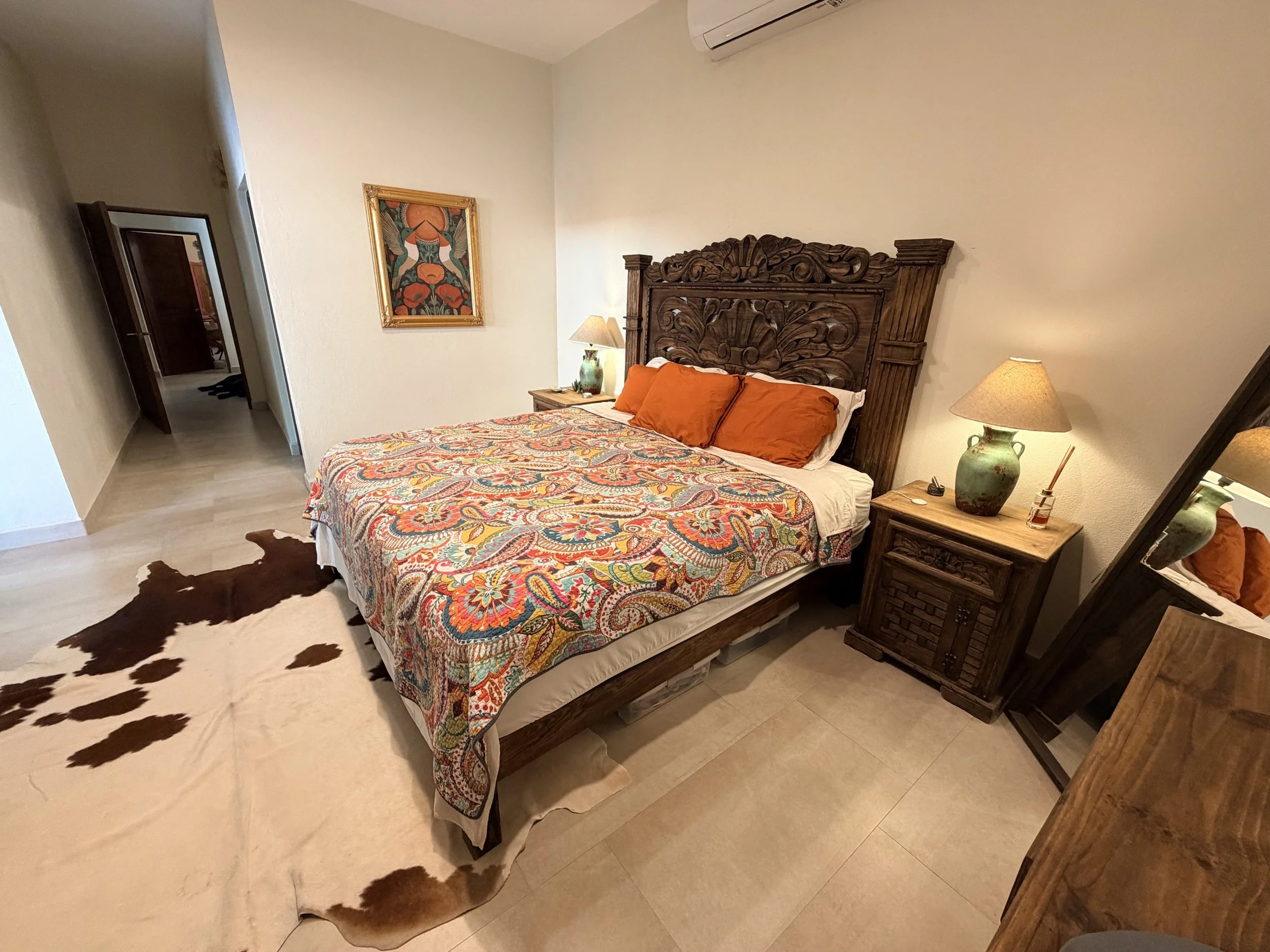 A bedroom with a large wooden bed with an intricate carved headboard. The bed has a colorful paisley-patterned bedspread and three orange pillows. There are two matching wooden nightstands, each with a lamp and decorative items. A cowhide rug is on t