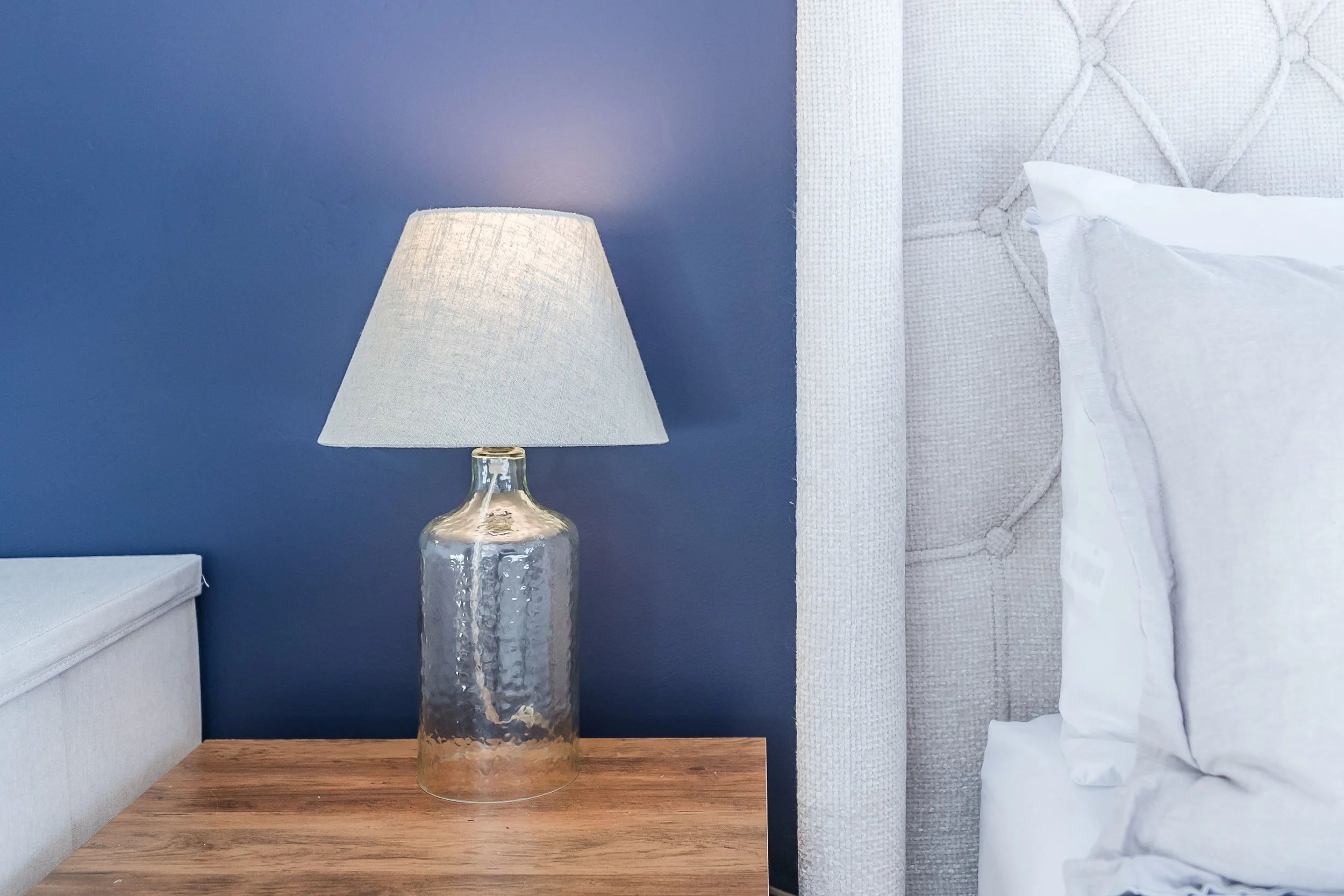 A bedroom corner featuring a dark blue wall with a table lamp that has a textured glass base and a light-colored lampshade. Part of a white upholstered bed with a tufted headboard and white bedding is visible on the right. A light-colored side table 