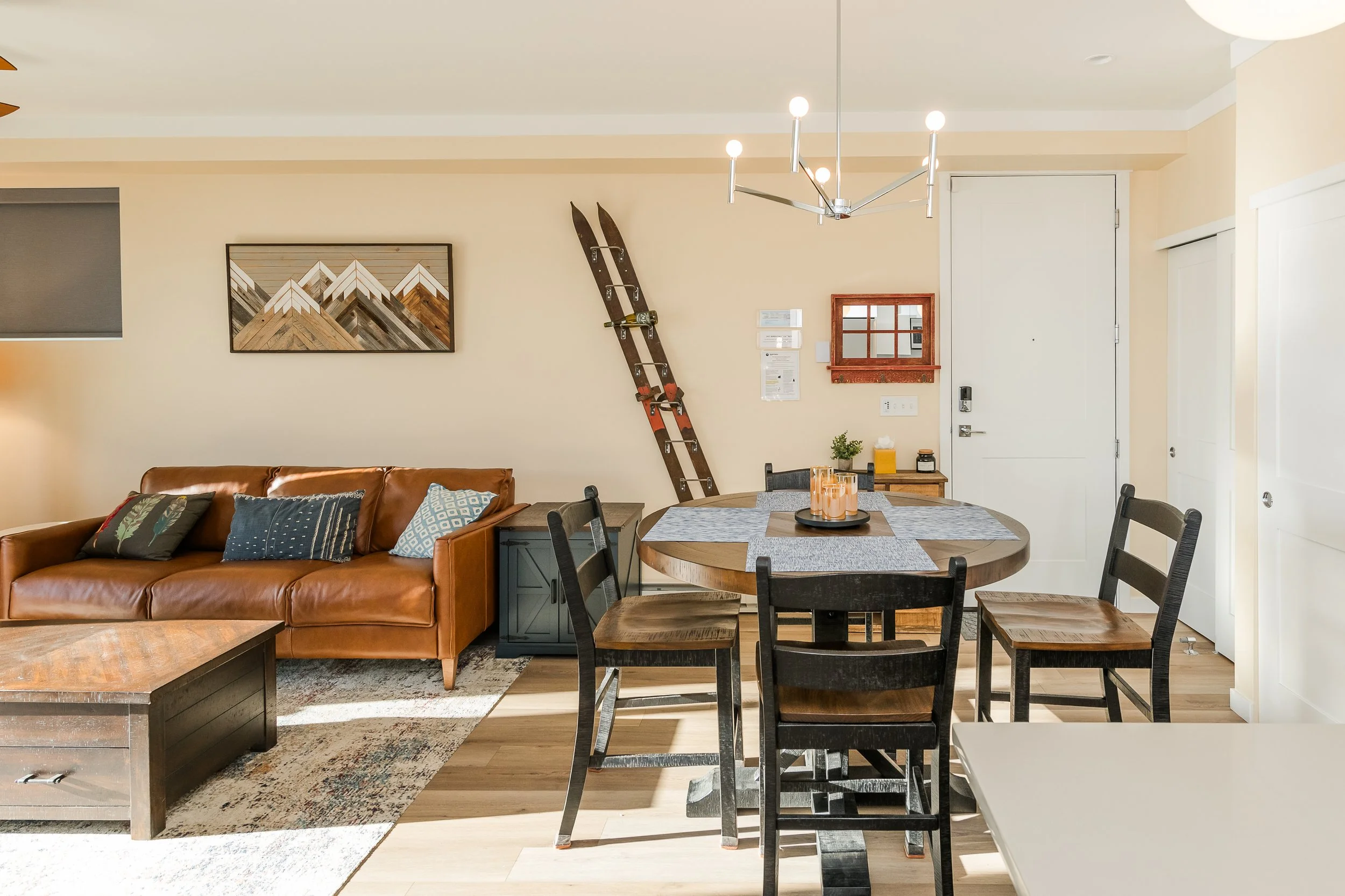 Interior of a living room with a brown leather couch, a round wooden dining table with black chairs, wall art, an old wooden skis, and various decorative items.