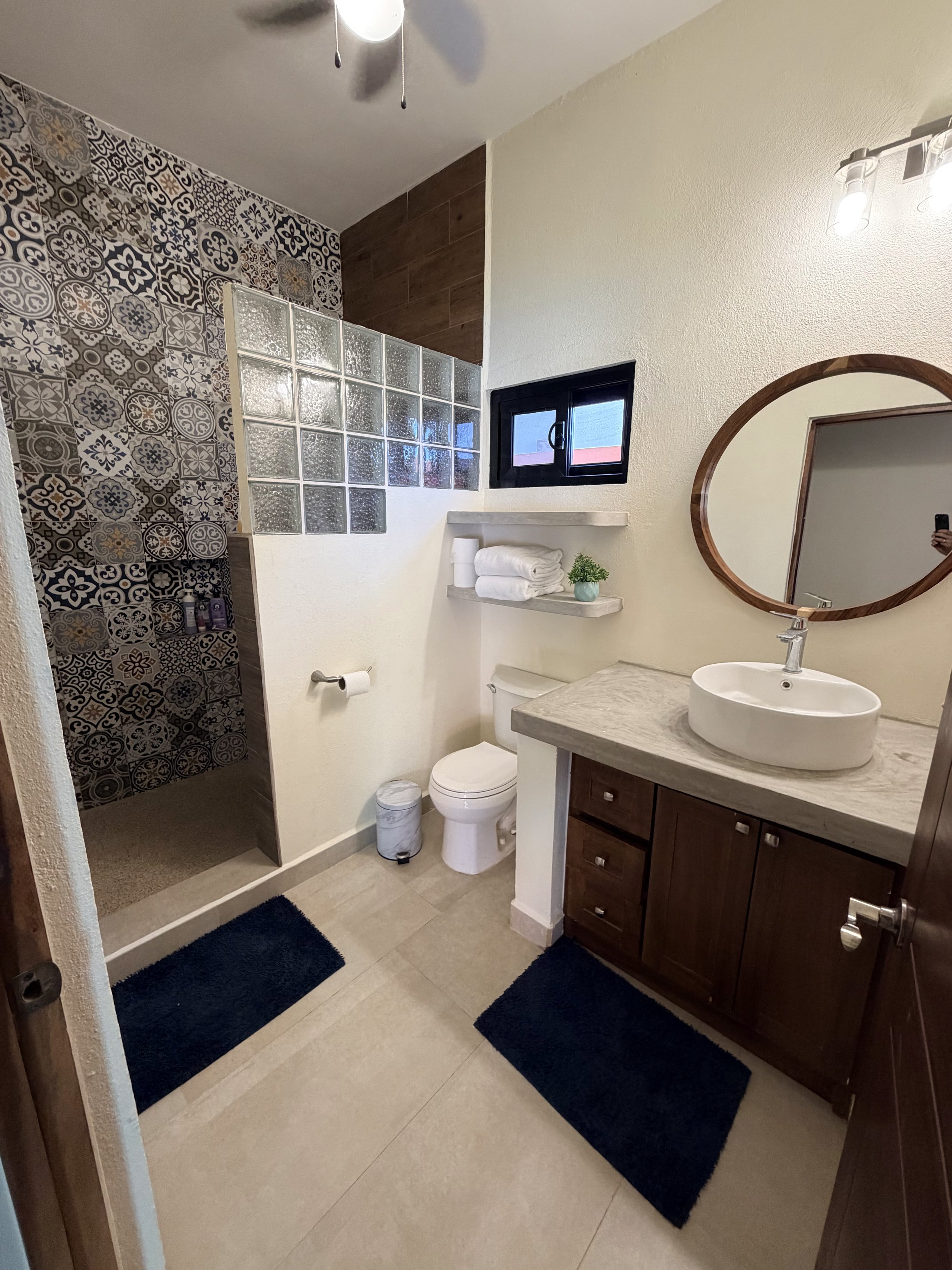 Bathroom with beige tiled floor, dark blue rugs, a white oval sink, wooden vanity with drawers, a mirror with a round wooden frame, a small window, white towels on shelves, a toilet, and a glass block partial wall separating the shower area with patt