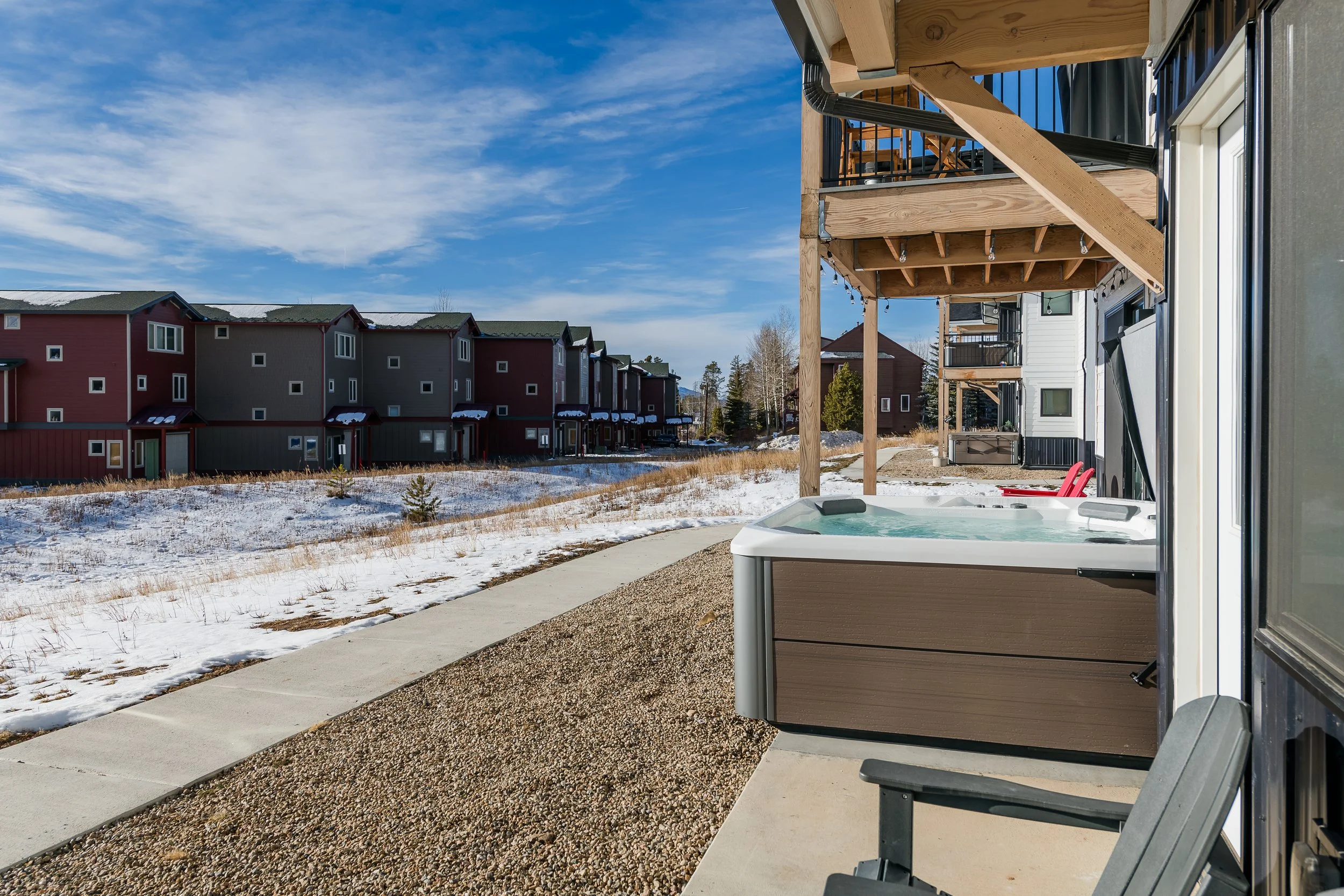 A shot of an apartment complex with a balcony and a hot tub outside under a wooden deck. The building is modern with black and white siding, and the area has snow on the ground with a clear blue sky.