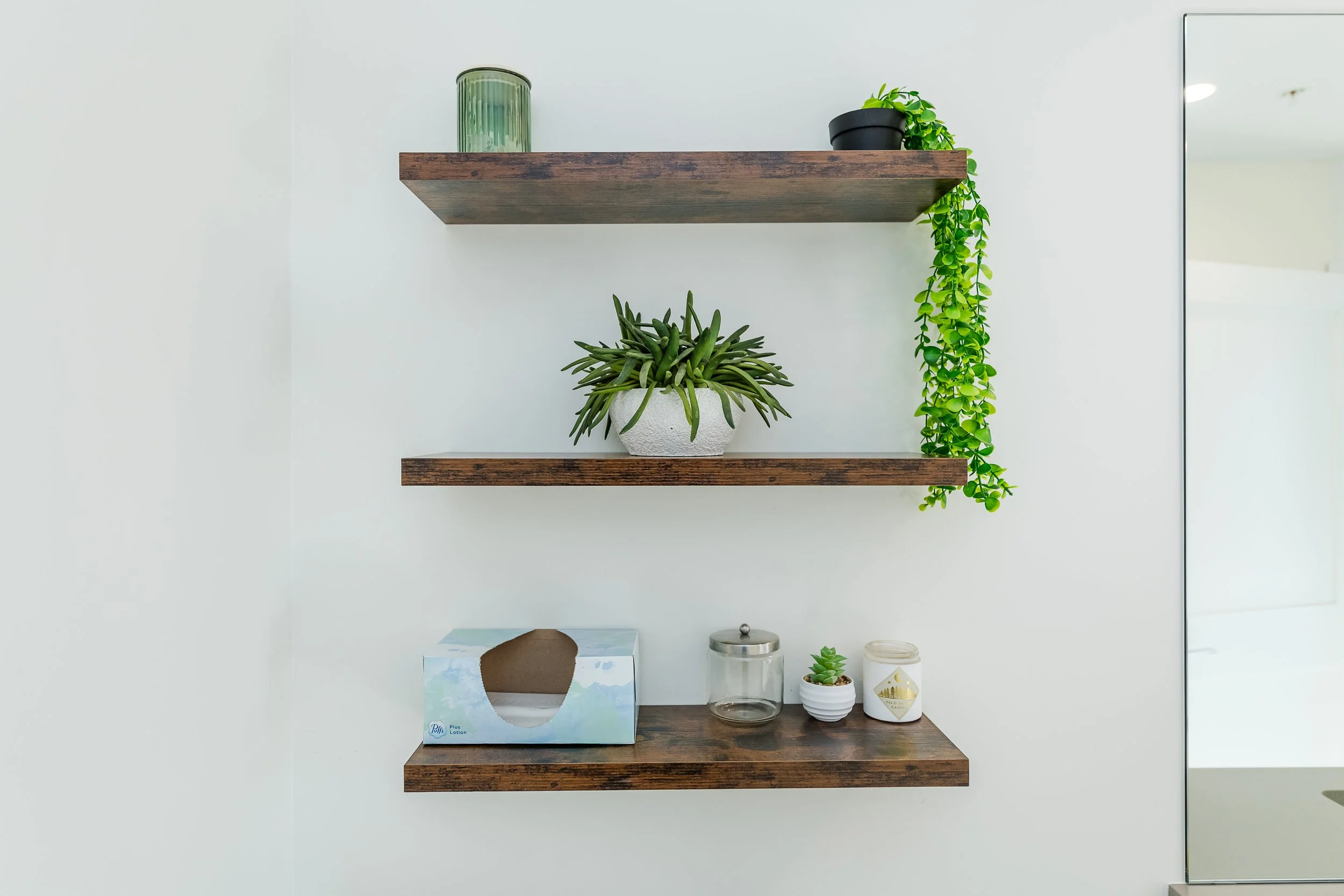 Two wooden floating shelves on a white wall with potted plants, glass jars, a tissue box, and candles.