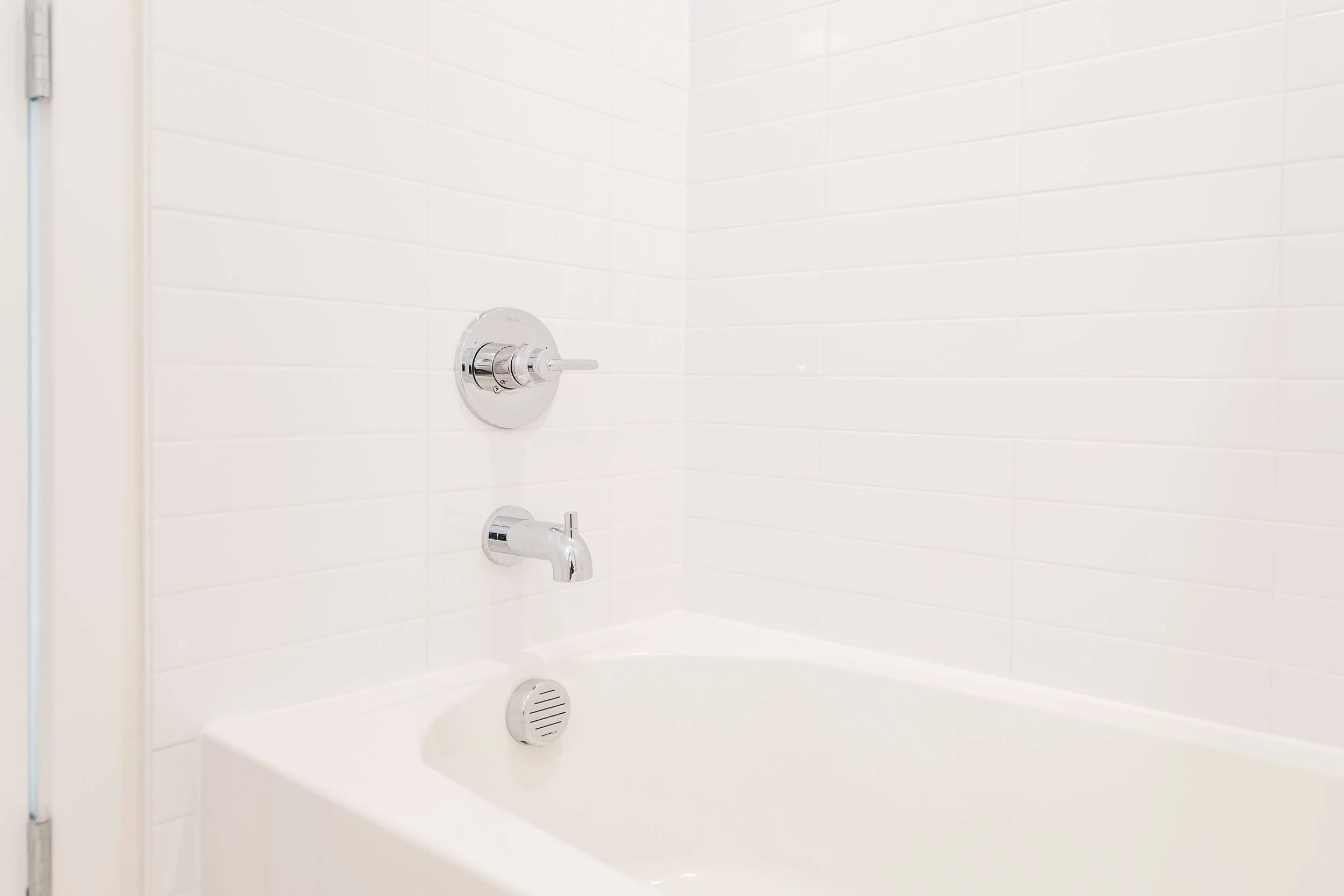 A white bathtub with a chrome faucet and fixtures, white tiled walls, and a drain at the bottom of the tub, in a modern bathroom.