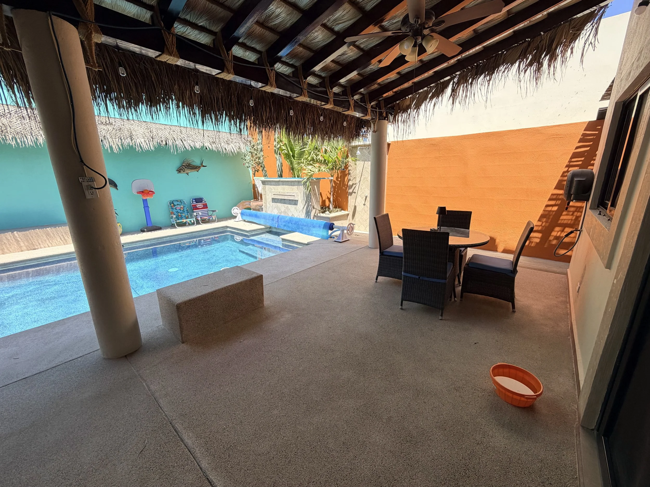 Covered outdoor patio area with a swimming pool, lounge chairs, and tropical plants, featuring a thatched roof and colorful walls.