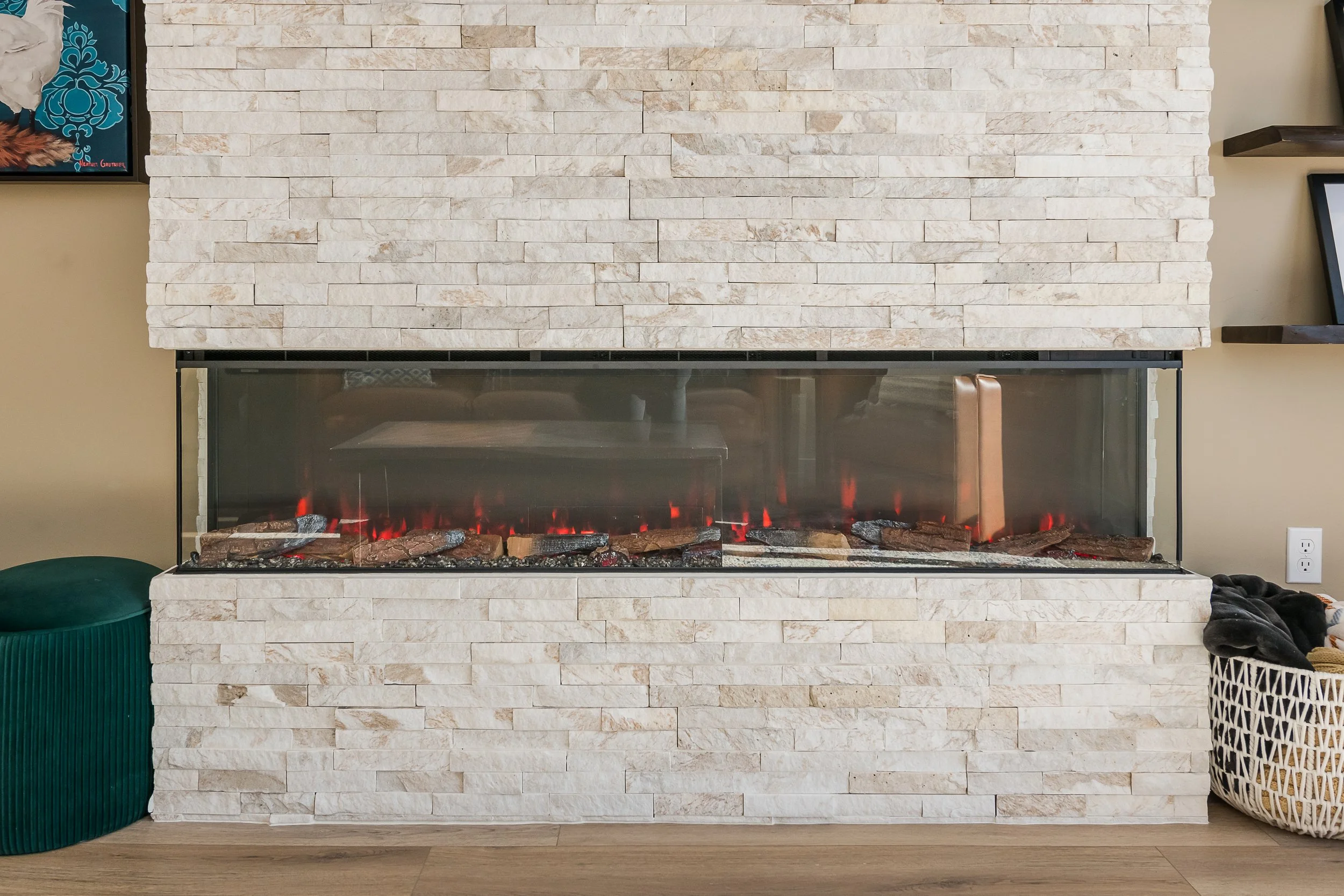 A fireplace with a white stone surround and a glass door, showing a simulated fire with glowing embers and logs inside. To the left is a green storage container, and to the right are woven baskets and a blanket.