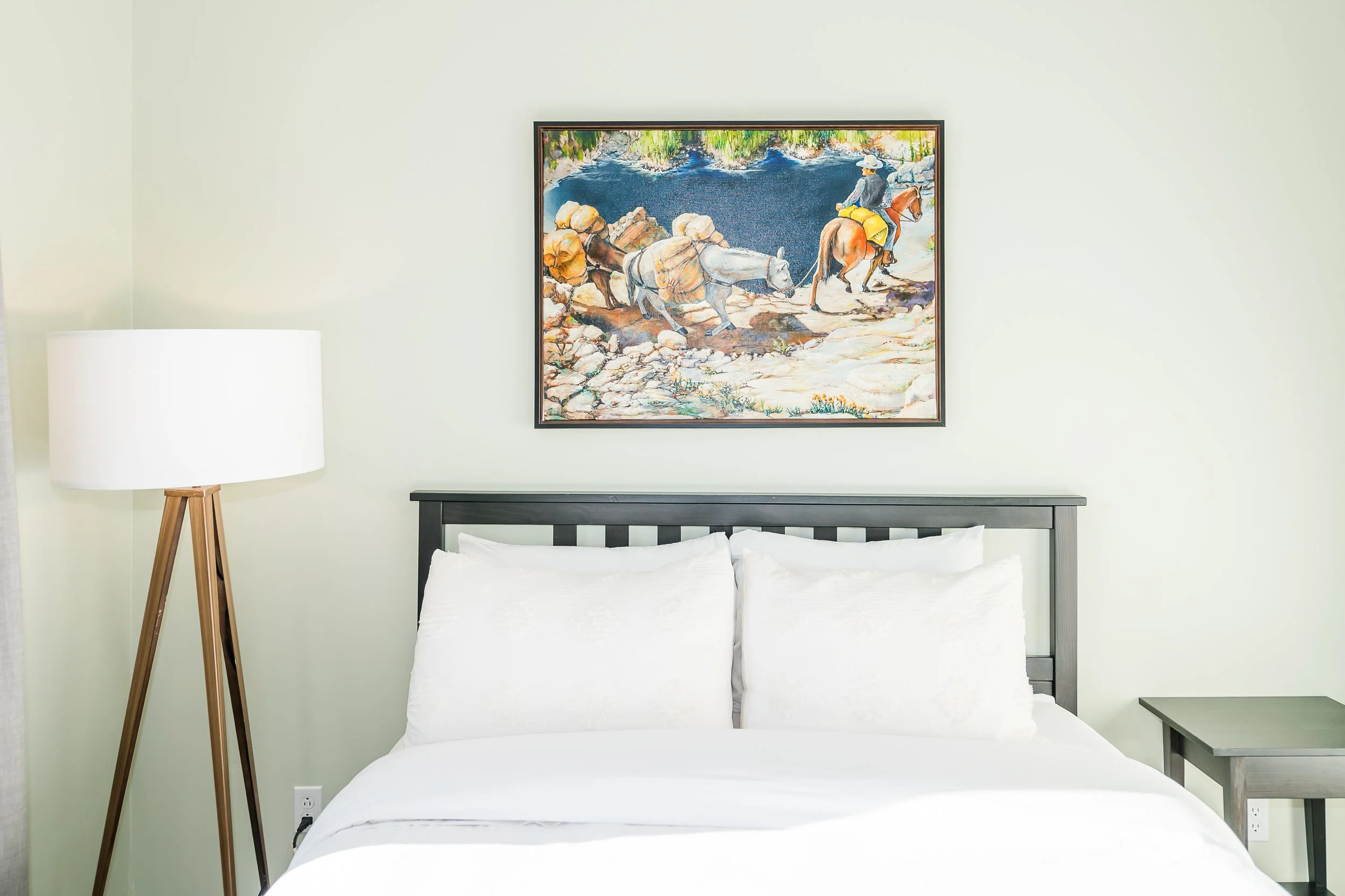 A bedroom with a bed with white bedding, a black bedside table, a floor lamp, and a framed picture of a mountain stream with people riding horses on the wall.
