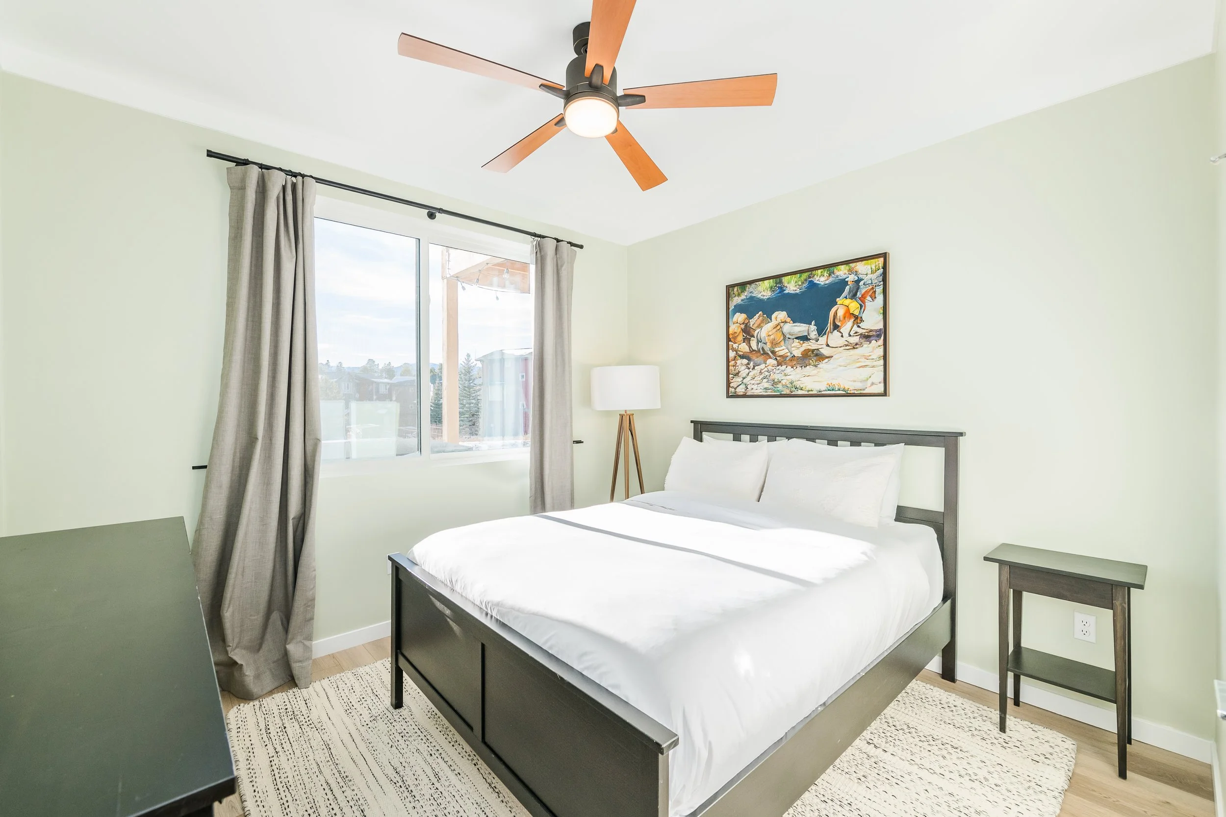 A bedroom with light green walls, a large window with grey curtains, a ceiling fan with wooden blades, a bed with white bedding, a wooden floor, a painting of animals, a white floor lamp, and a small black side table.