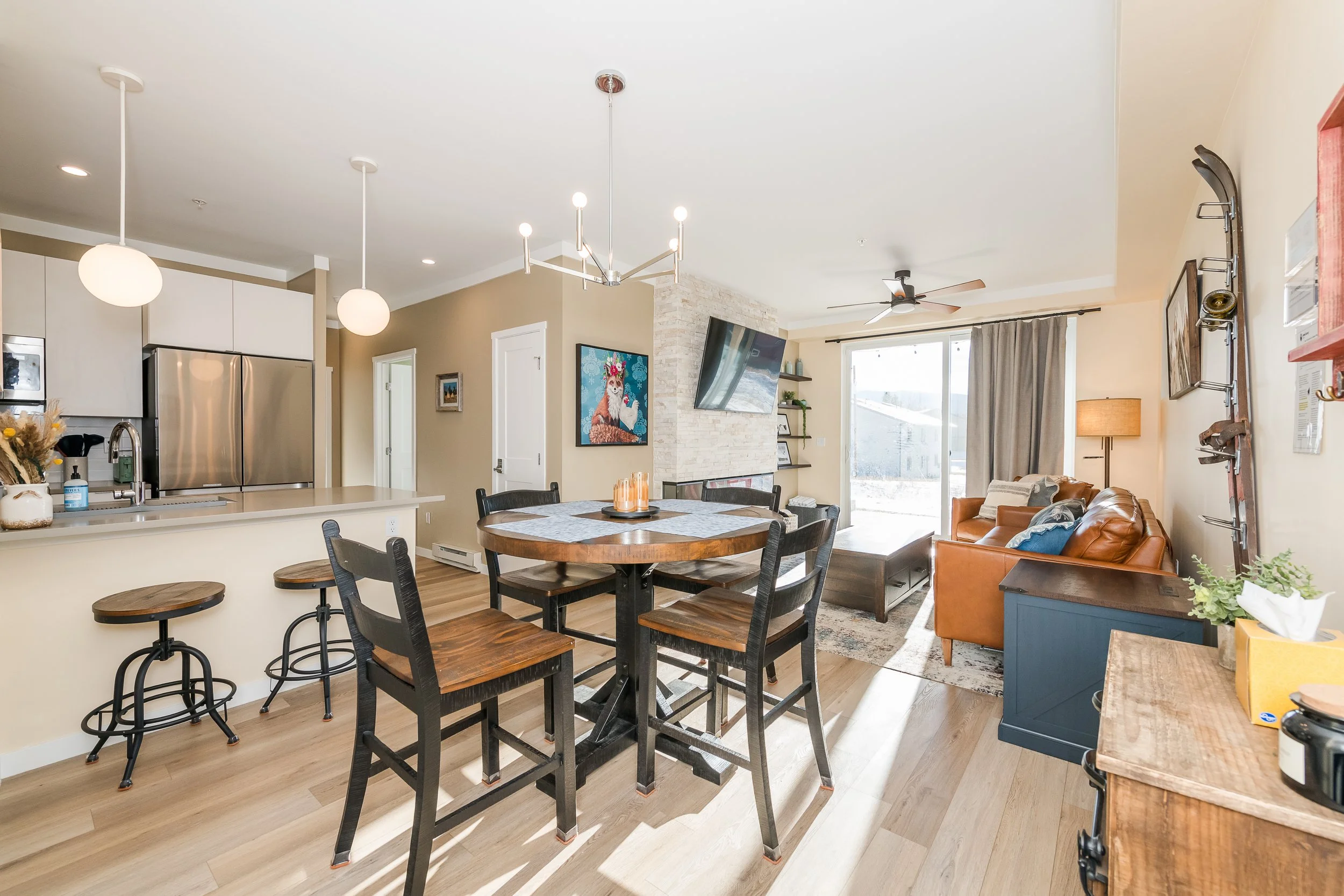 Open-concept living space with kitchen, dining, and living areas, featuring pendant lights, a ceiling fan, and a sliding glass door with beige curtains.