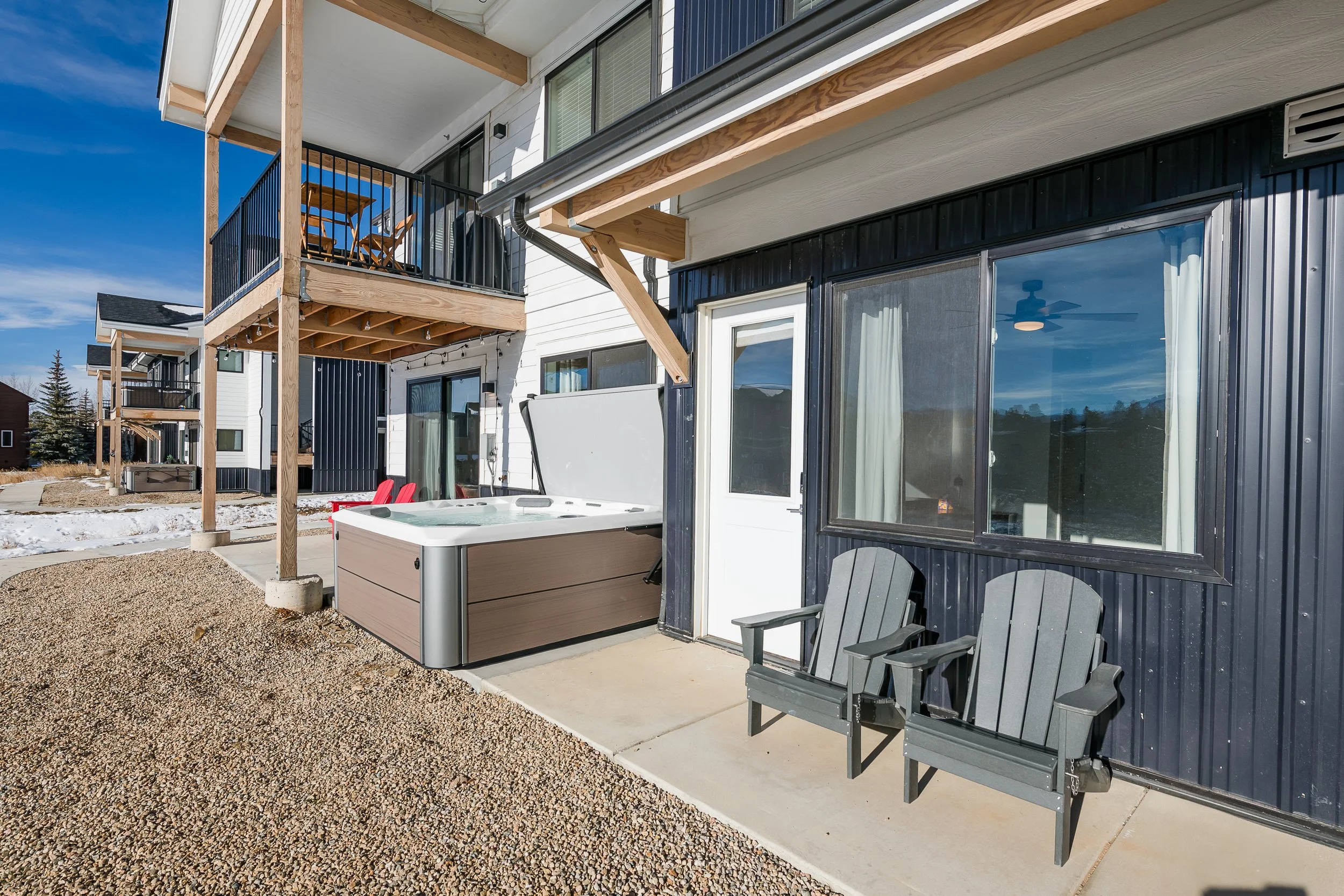 Back patio of a modern apartment building with a balcony, outdoor chairs, hot tub, and view of neighboring houses and blue sky.