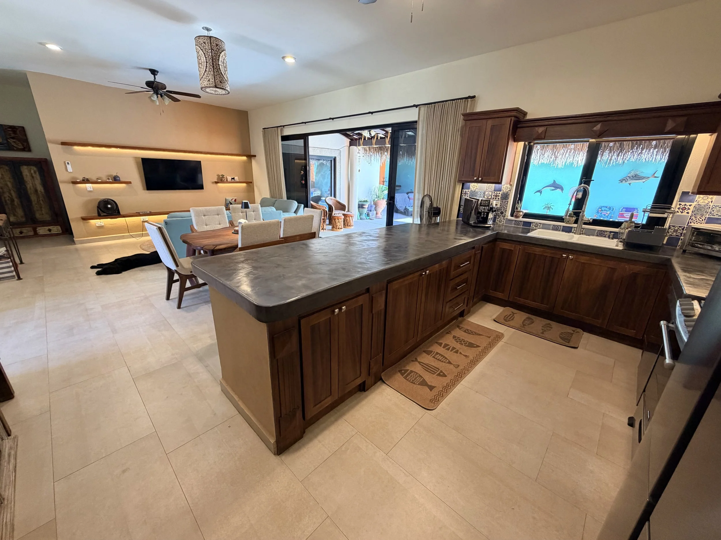 Open-concept living and kitchen area with sliding glass door leading to outdoor patio, with frosted windows featuring dolphin images, wooden cabinetry, beige tiled floor, and a dining table with six chairs.