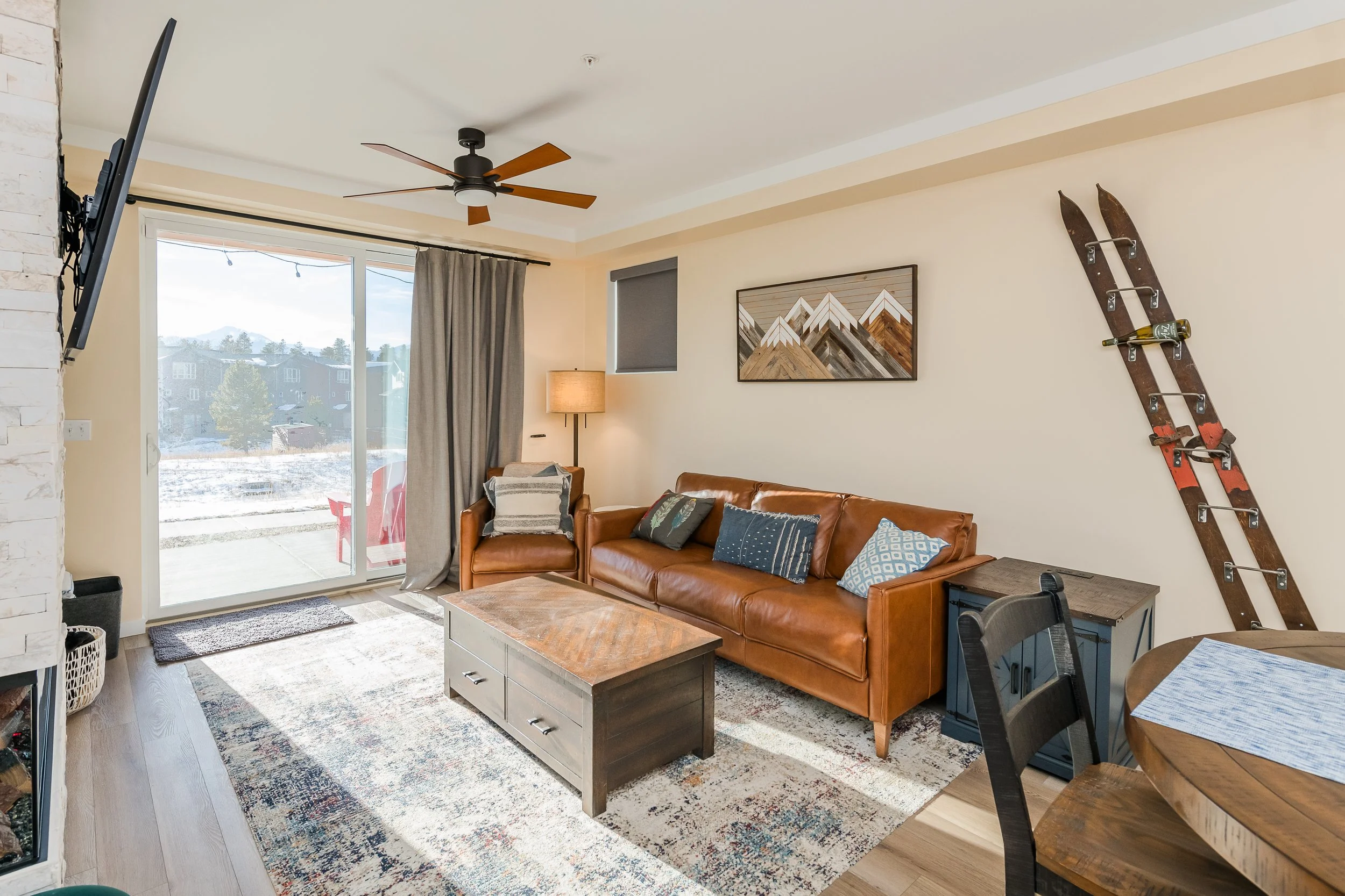 Living room with large sliding glass door showing snowy outdoor scenery, a leather sofa with decorative pillows, a wooden coffee table, a mounted television, a rustic wall art, and a vintage wooden ski with a wine bottle on it.