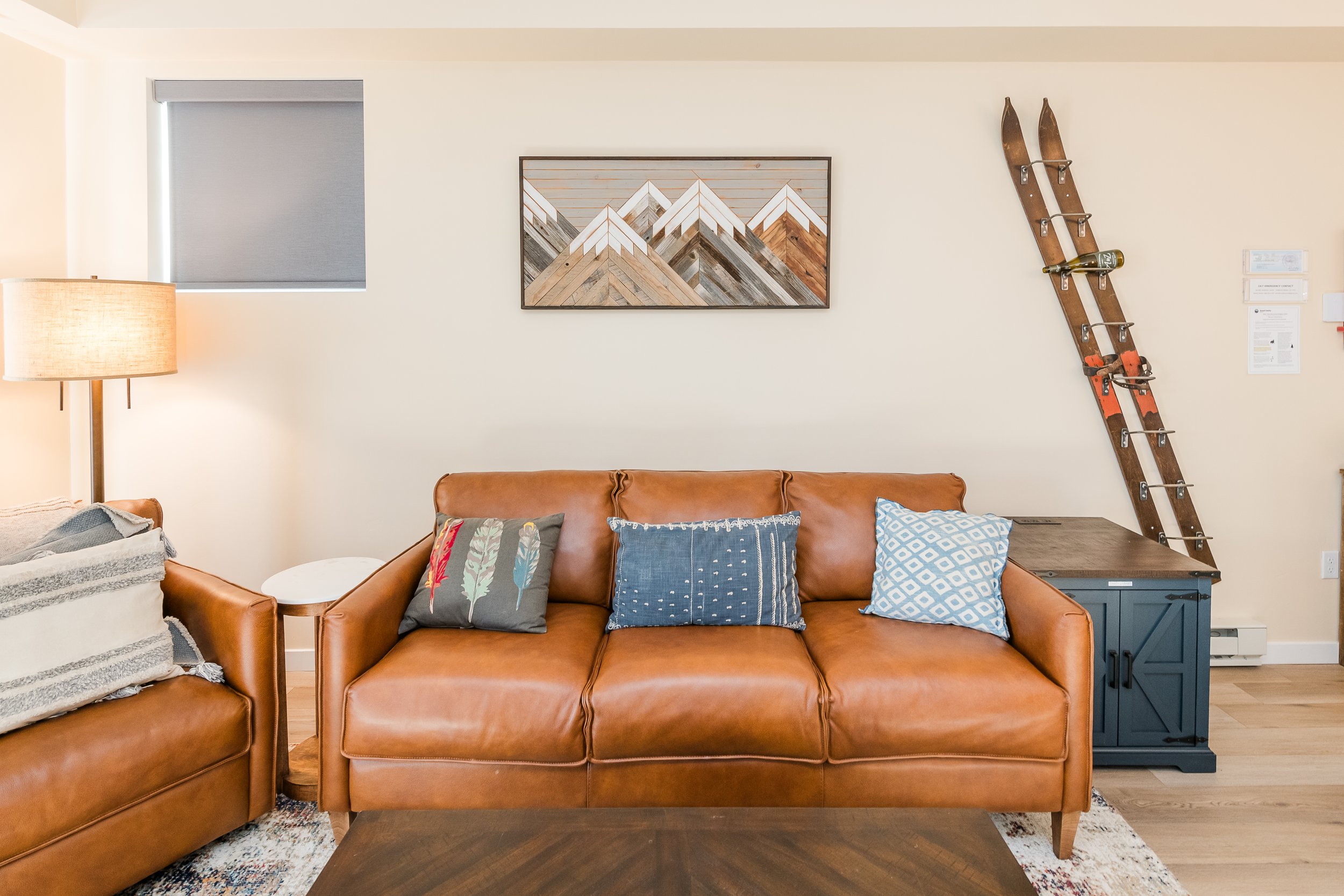 Living room with brown leather sofa, decorative pillows, wooden side table, floor lamp, mountain artwork, vintage wooden skis on wall, window with closed blinds, light-colored walls.