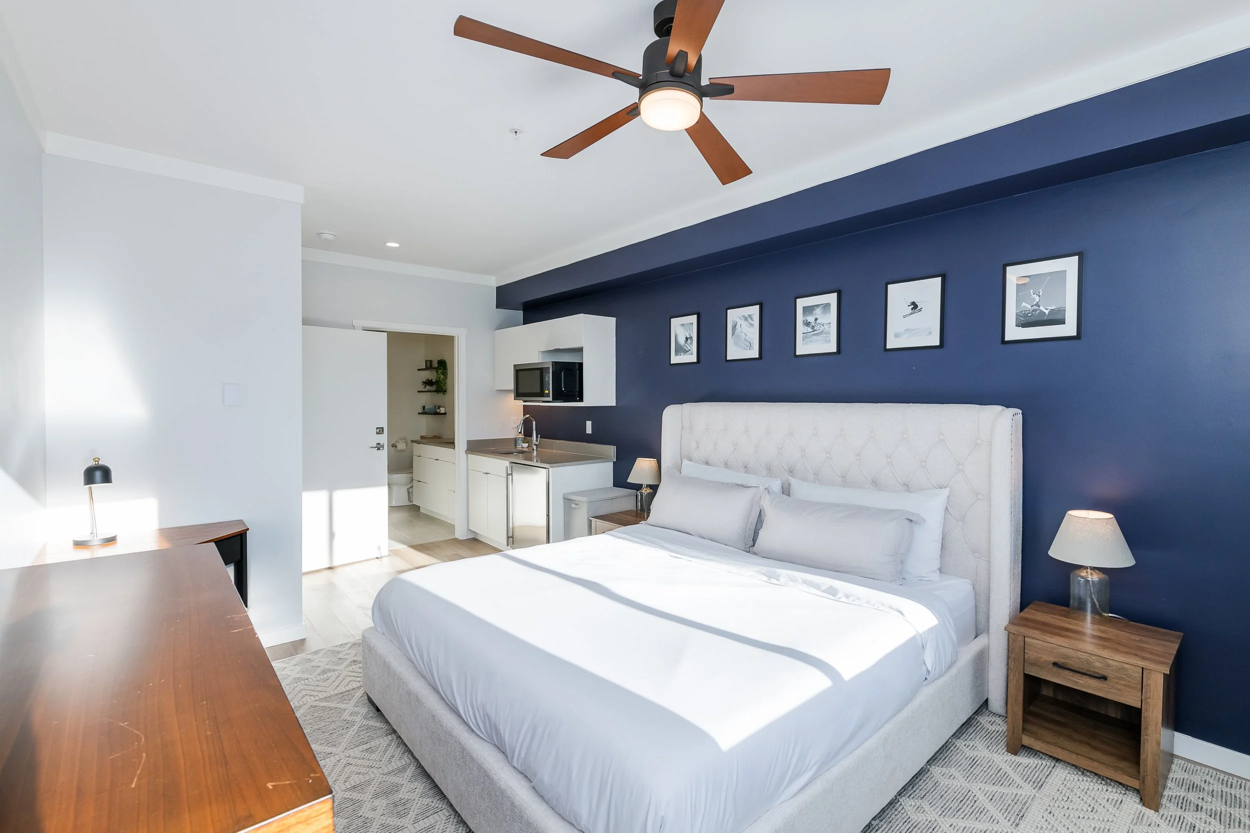 A hotel room with a white bed, bedside tables, lamps, and a navy blue accent wall with framed black-and-white pictures of surfers. The room has a ceiling fan with wooden blades and a central light. There is a small kitchenette area with a microwave, 