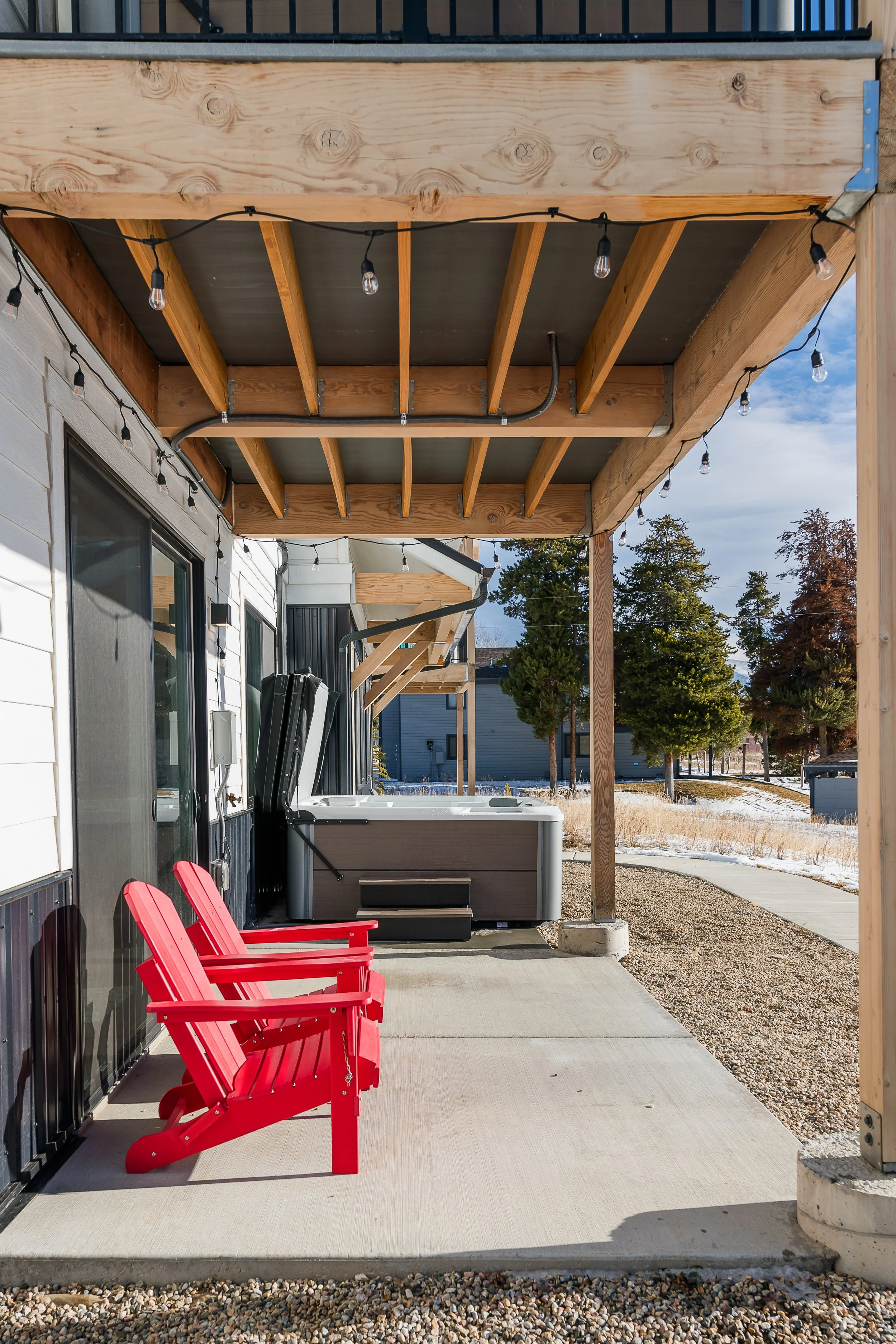 A covered outdoor patio area with two red Adirondack chairs, wood beams, string lights, a hot tub, and trees in the background.