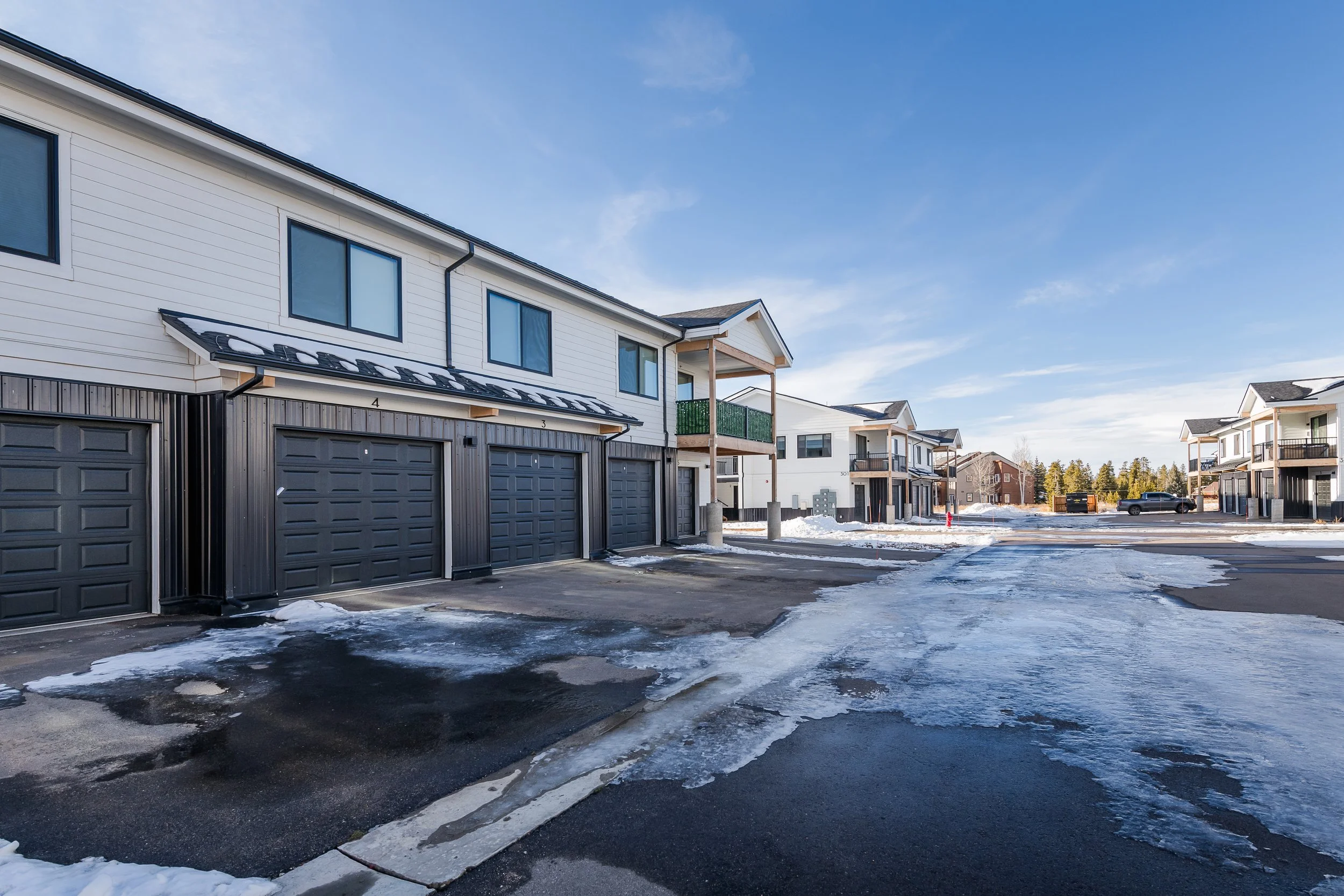 A row of modern townhouses with garages in a snowy neighborhood under a clear blue sky.
