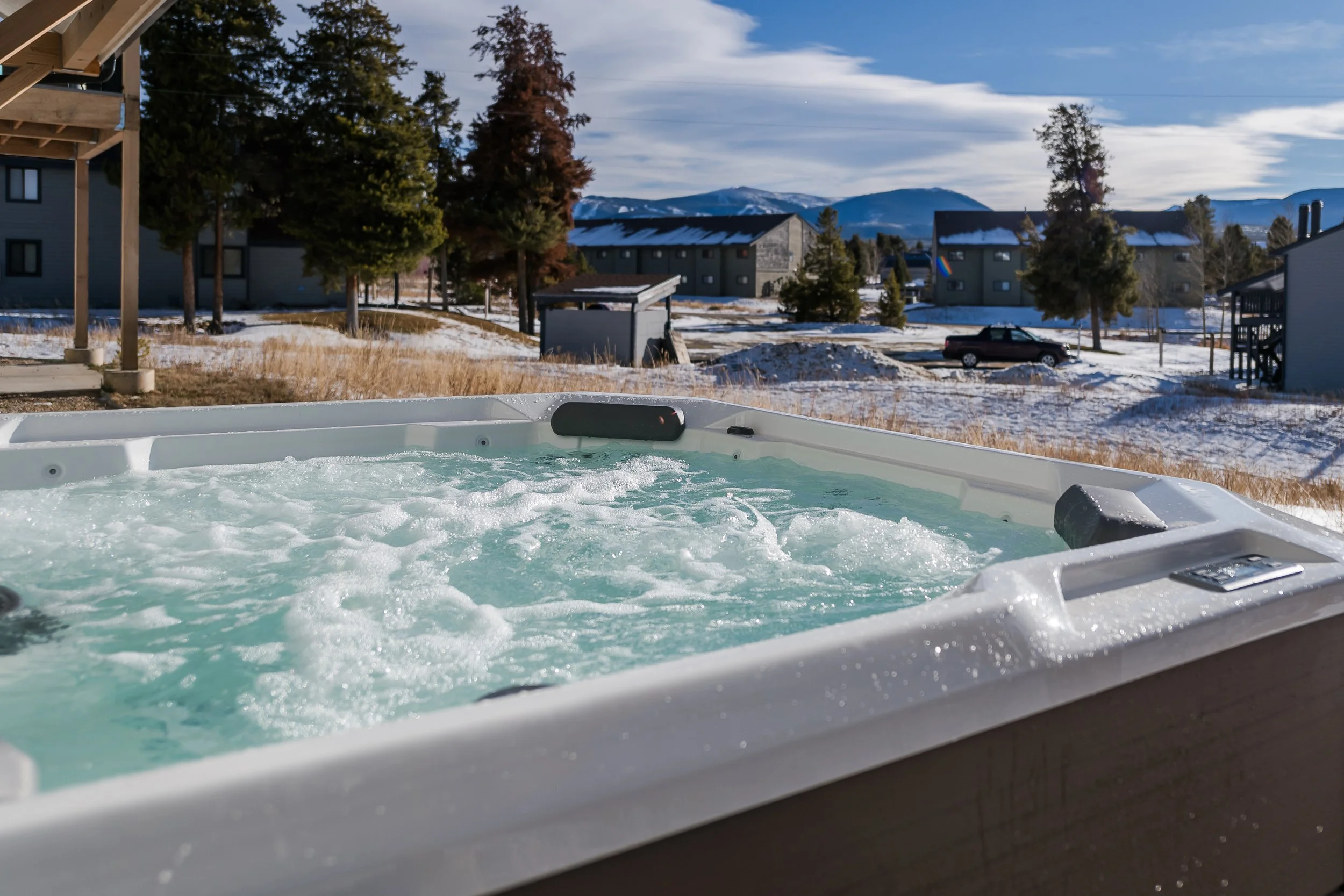 View from a hot tub overlooking snowy residential neighborhood with trees, houses, a black vehicle, and mountains in the background on a partly cloudy day.