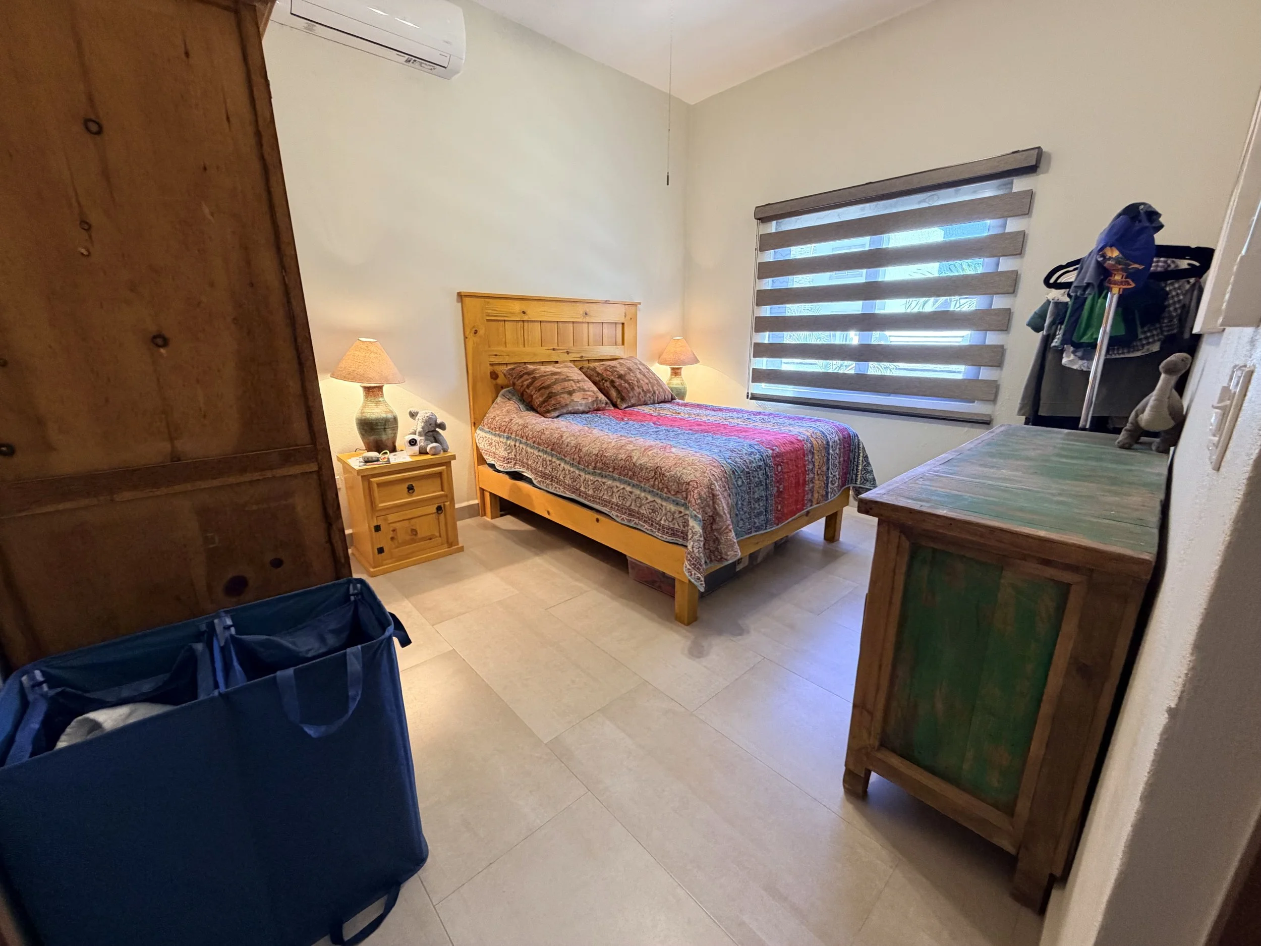 A bedroom with a wooden bed, colorful quilt, and two bedside lamps. There is a window with horizontal blinds. A wooden dresser and a clothing rack with clothes are visible. In the foreground, there are blue bags on the floor.