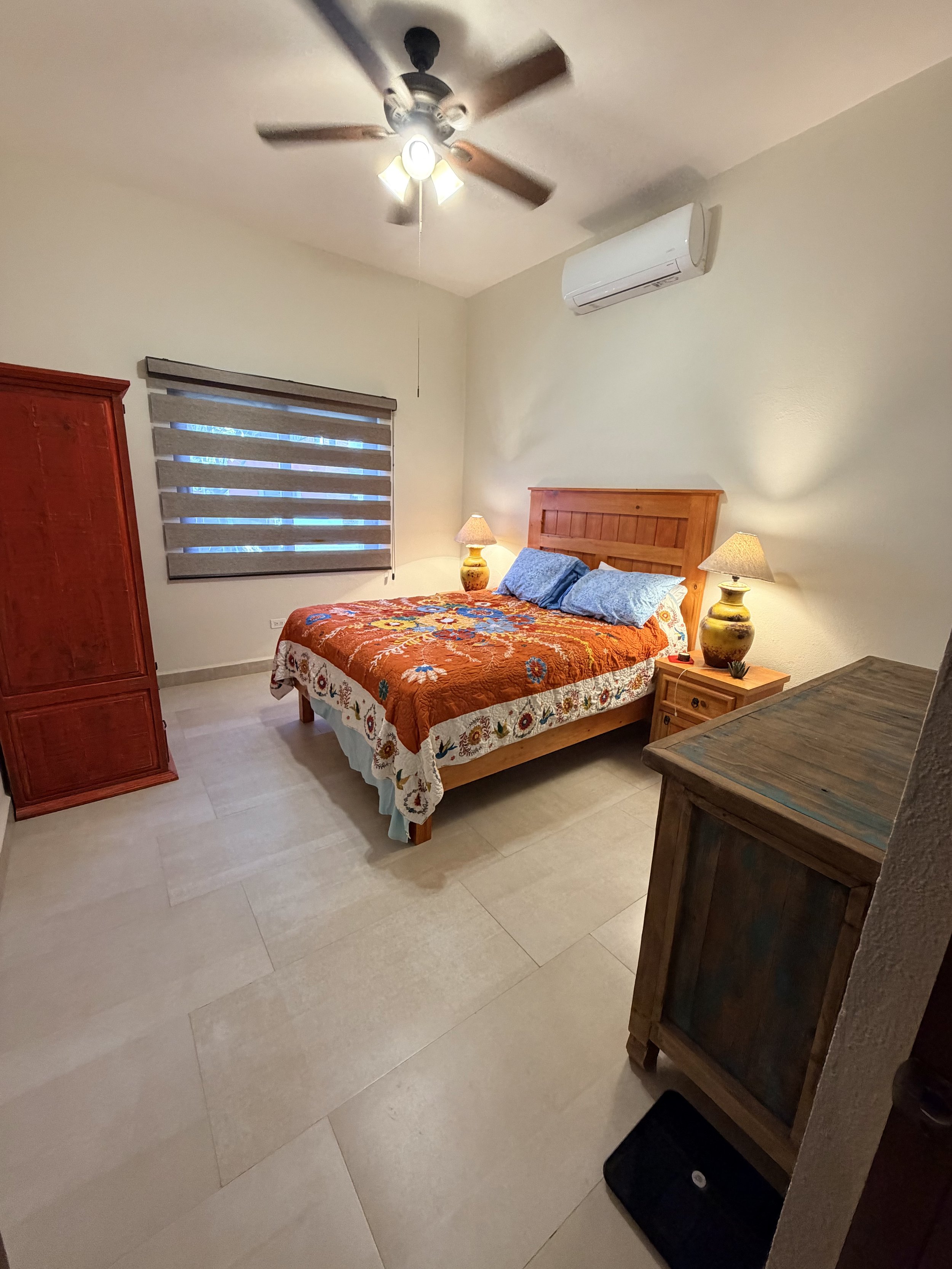 A bedroom with a wooden bed, colorful bedding, two lamps on bedside tables, a window with gray blinds, a wooden wardrobe, a ceiling fan, an air conditioning unit, and tiled floor.