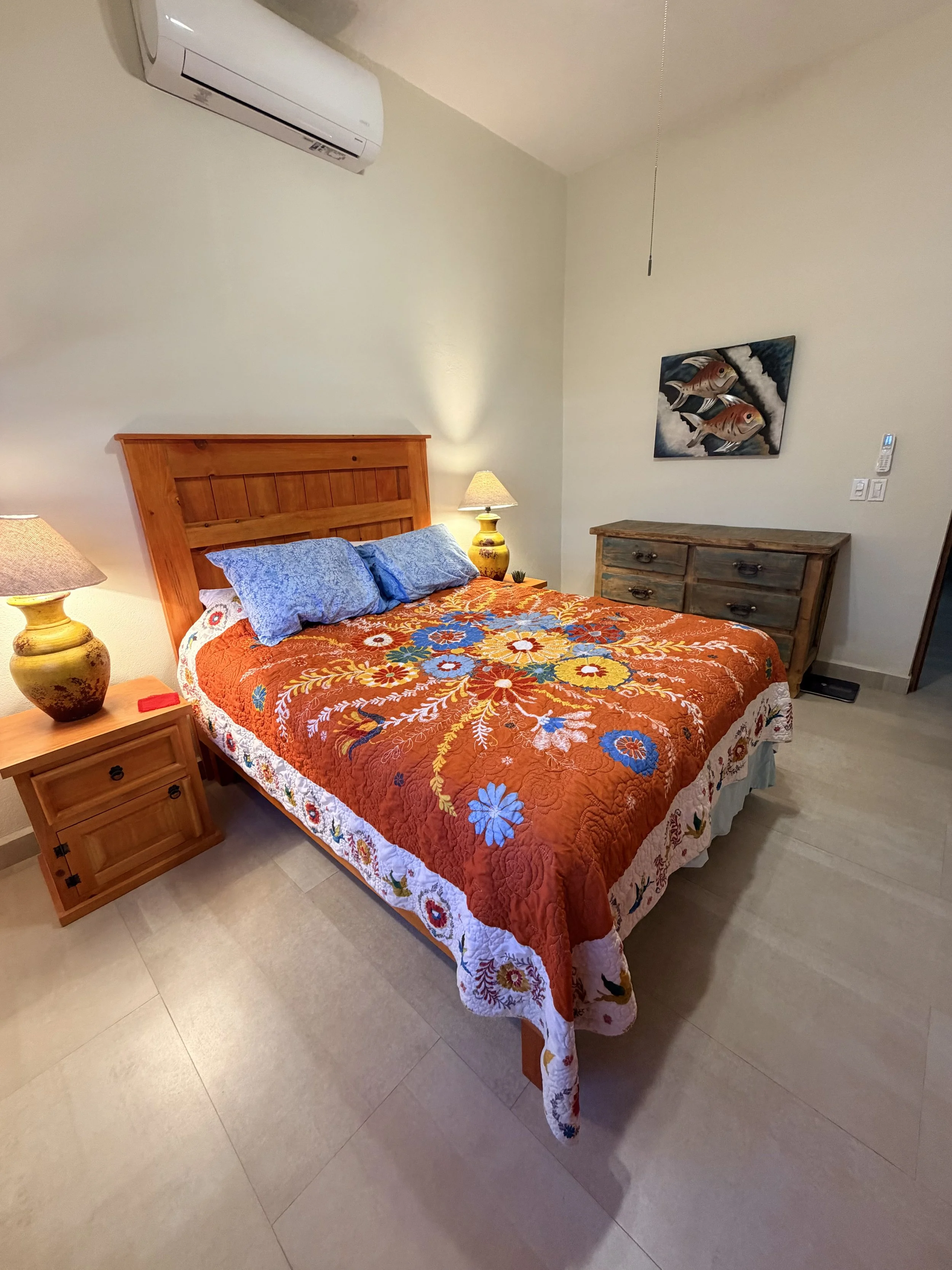 A bedroom with a wooden bed, colorful floral quilt, blue pillows, two yellow bedside lamps, a rustic wooden dresser, a wall-mounted painting of fish, and an air conditioning unit on the wall.