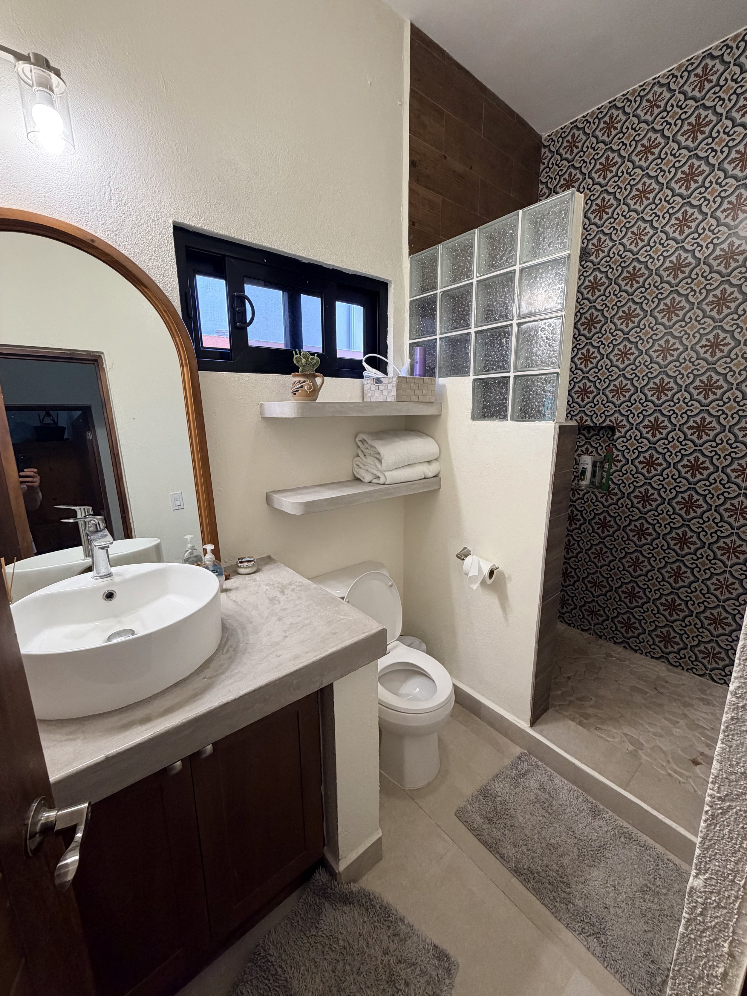 A small bathroom with a beige countertop, a round white vessel sink, a wooden framed mirror, a small window, open shelves with folded towels, and a shower area with patterned tiles and glass block wall.