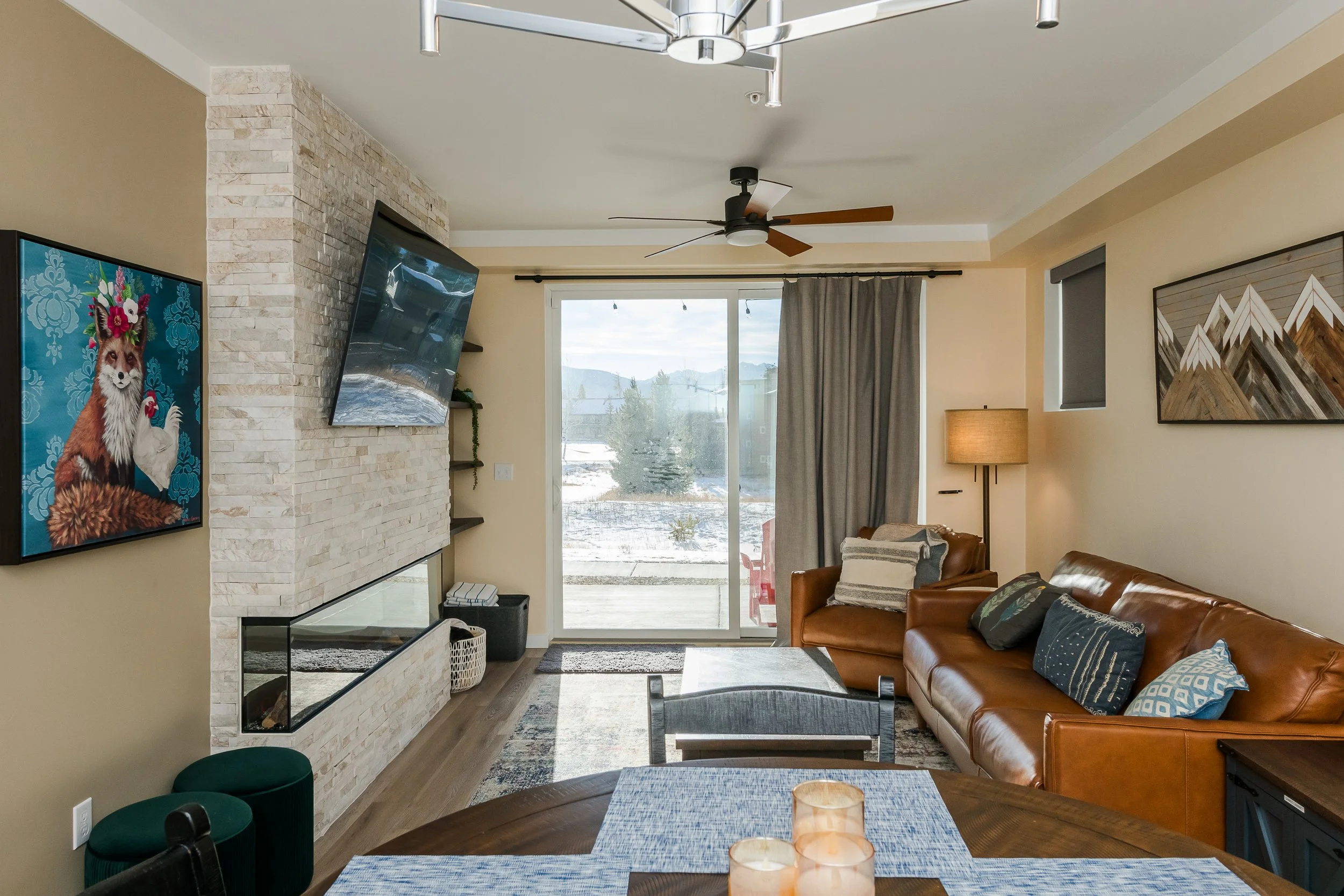 A living room with a beige wall, artwork, a brown leather sofa, a glass-top coffee table with candles, a sliding glass door leading outside, and a ceiling fan.