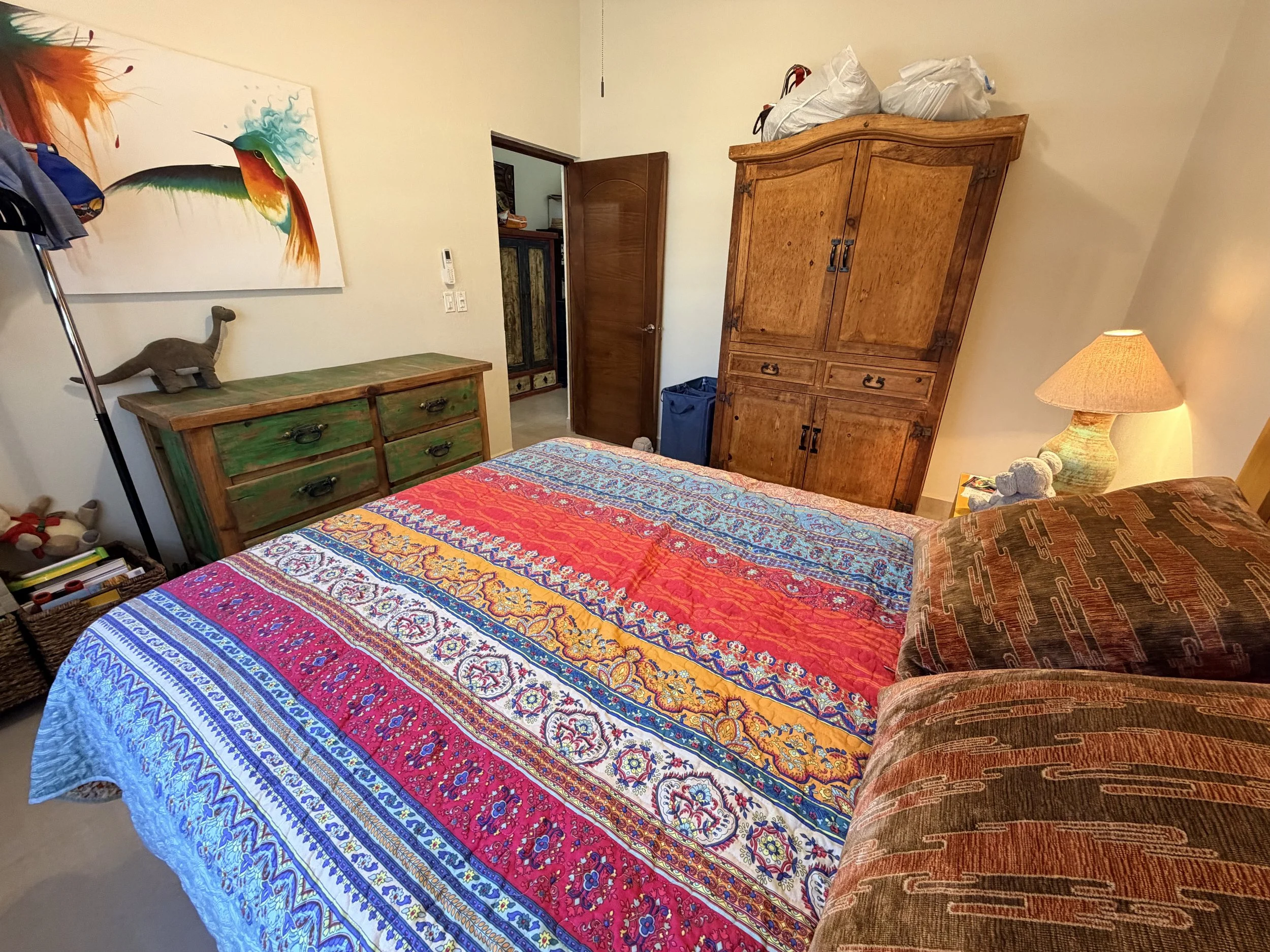 Bedroom with colorful quilted bedspread, a wooden dresser with a dinosaur plush toy, a large wooden wardrobe, a lamp, and artwork of a hummingbird on the wall.