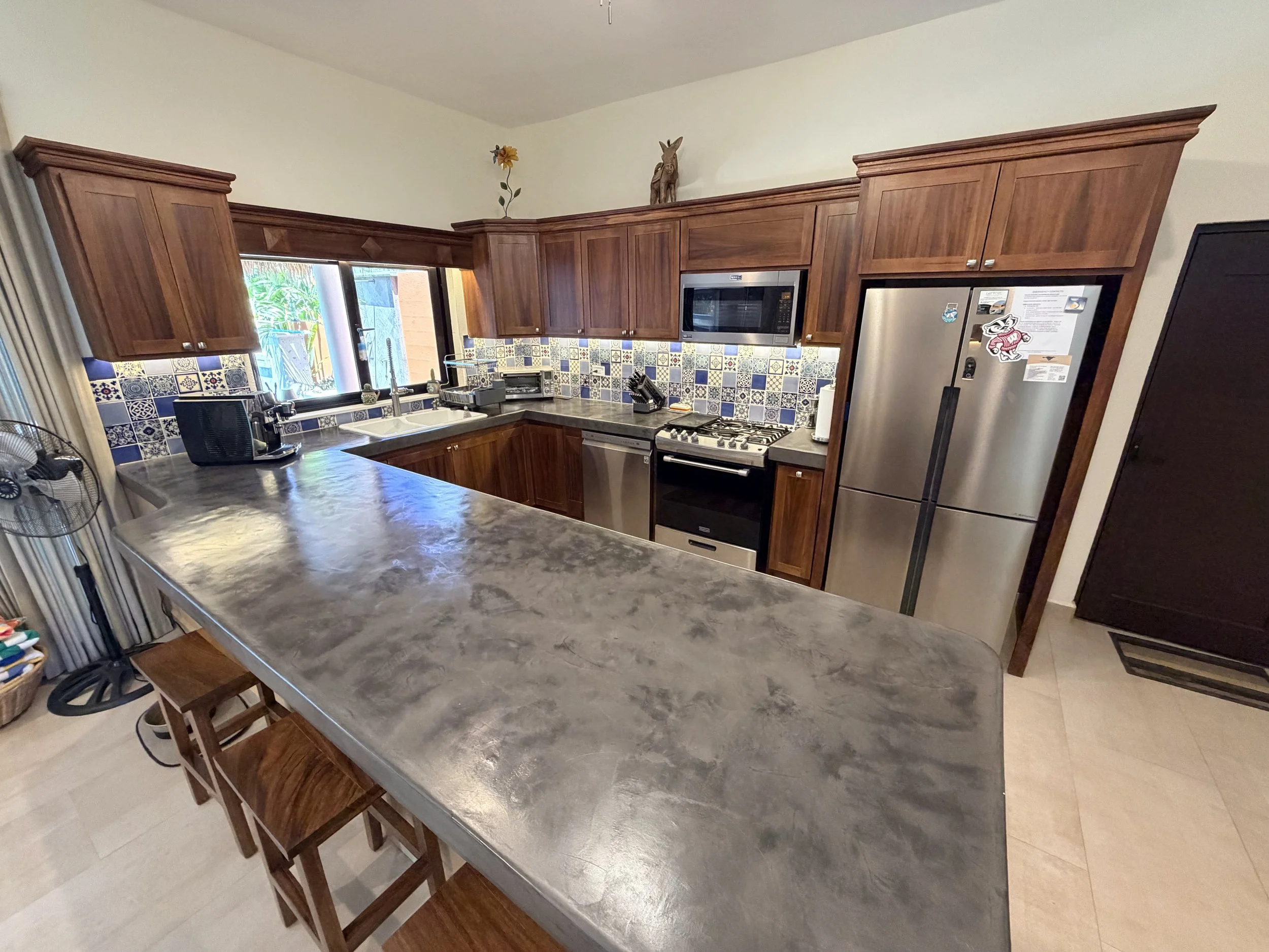A kitchen with wooden cabinets, a gas stove, a microwave, a refrigerator, and a countertop with various appliances and items. There is a window above the sink, and decorative tiles on the backsplash.