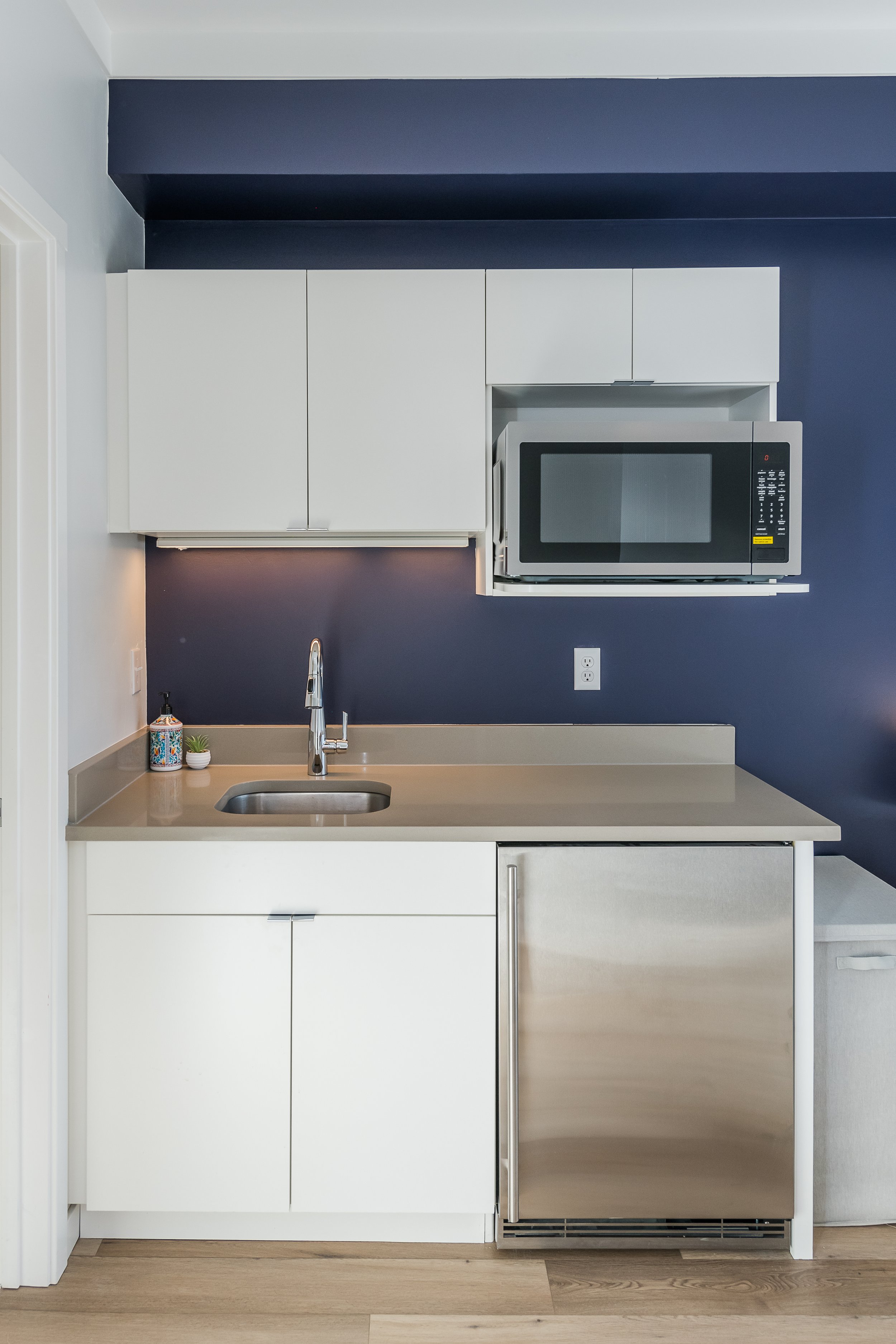 Modern kitchen with white cabinets, a stainless steel microwave, a beige countertop, a small potted plant, soap dispenser, a kitchen faucet, and a dark blue wall.