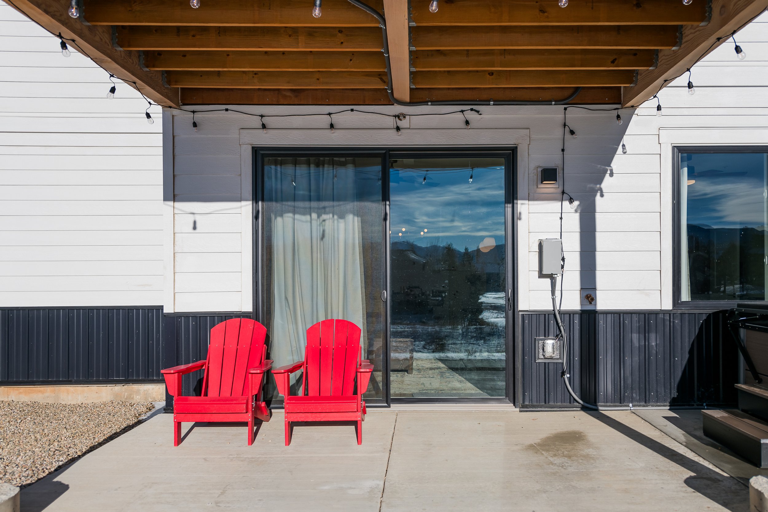 A backyard patio with two red Adirondack chairs on a concrete slab, a sliding glass door, and a white house exterior with black trim. String lights are hung above the patio, and there is a small covered balcony with a wooden underside above the chair