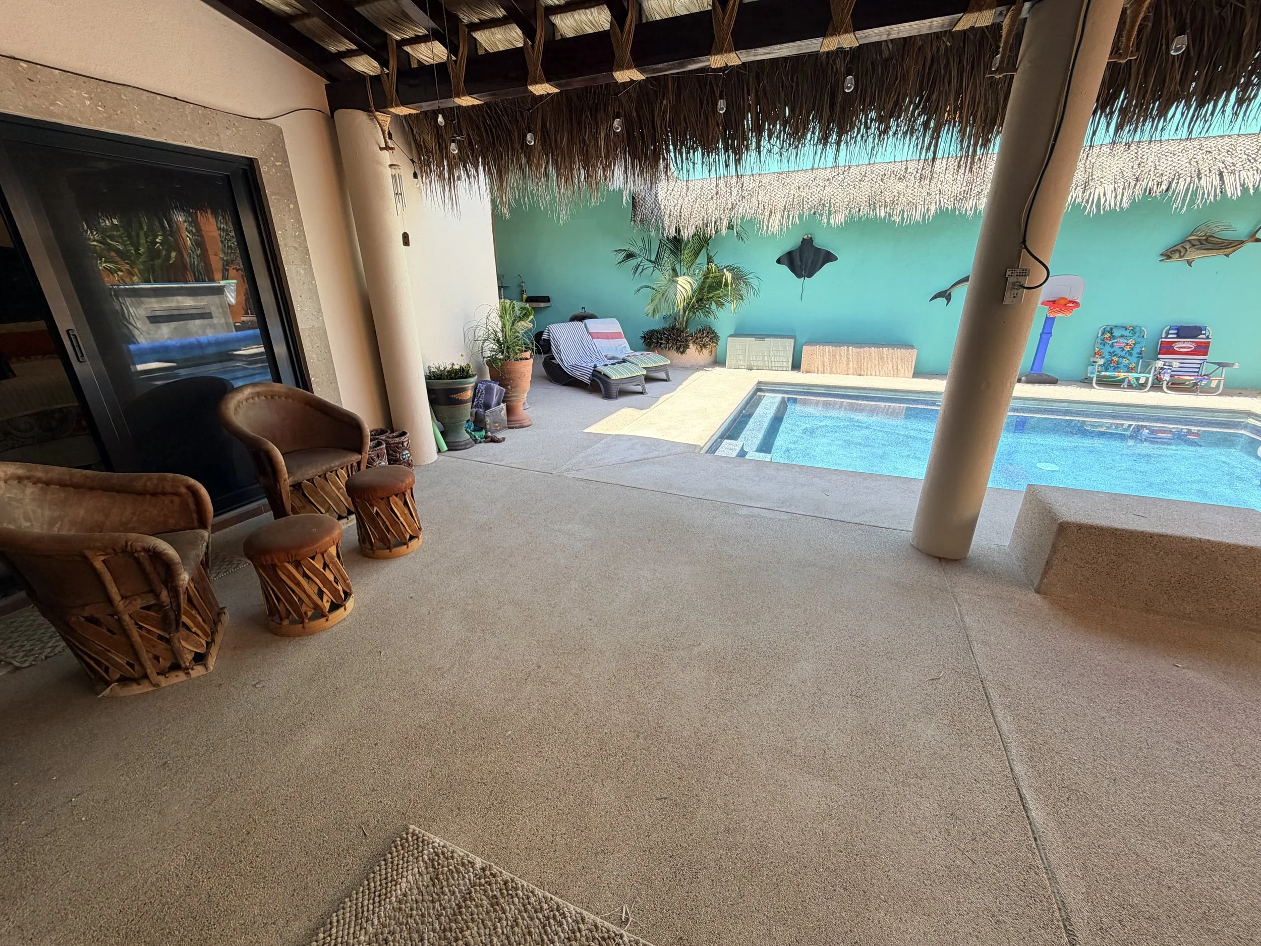 A covered outdoor patio area overlooking a swimming pool, with potted plants, lounge chairs, and decorative wall art of fish and a stingray.