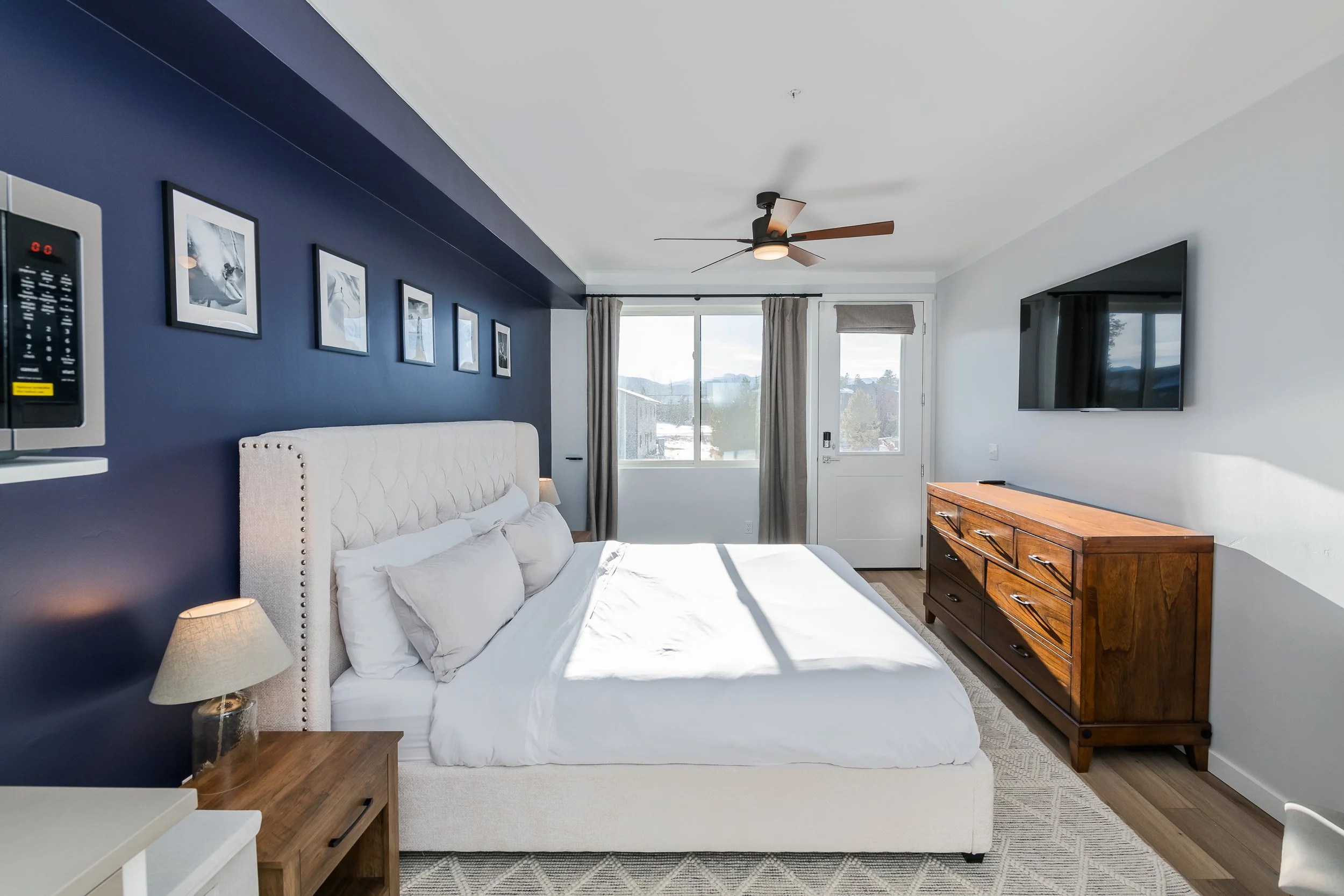 A bedroom with a white bed, a dark blue accent wall with framed black and white photos, a wooden dresser, a wall-mounted TV, a ceiling fan, and a sliding glass door with curtains leading outside.